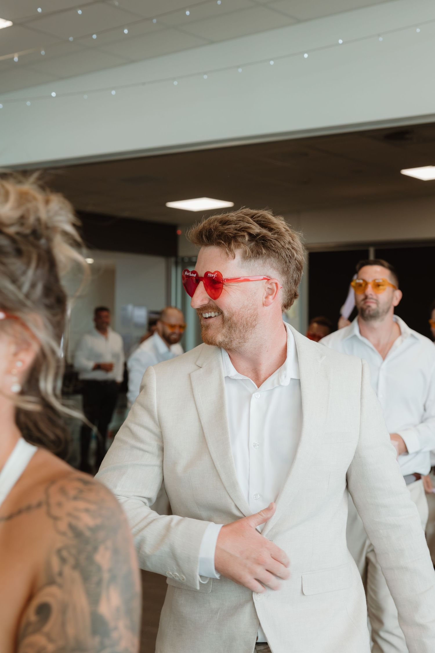 Man in beige suit wearing red heart-shaped sunglasses smiling at a wedding reception event.