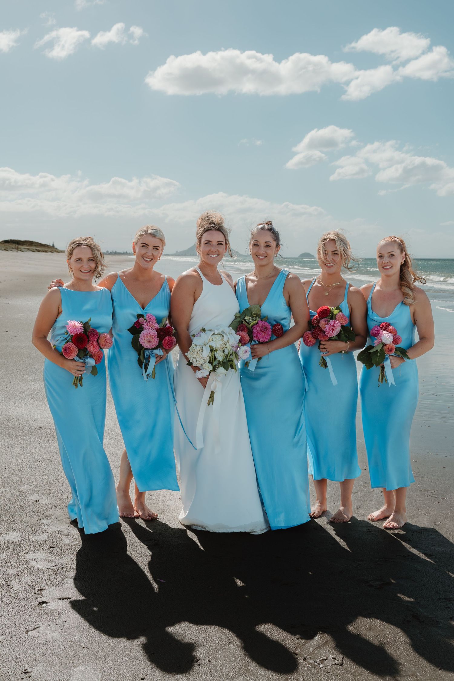 Bride in white gown with five bridesmaids in blue dresses holding bouquets on a sunny beach.