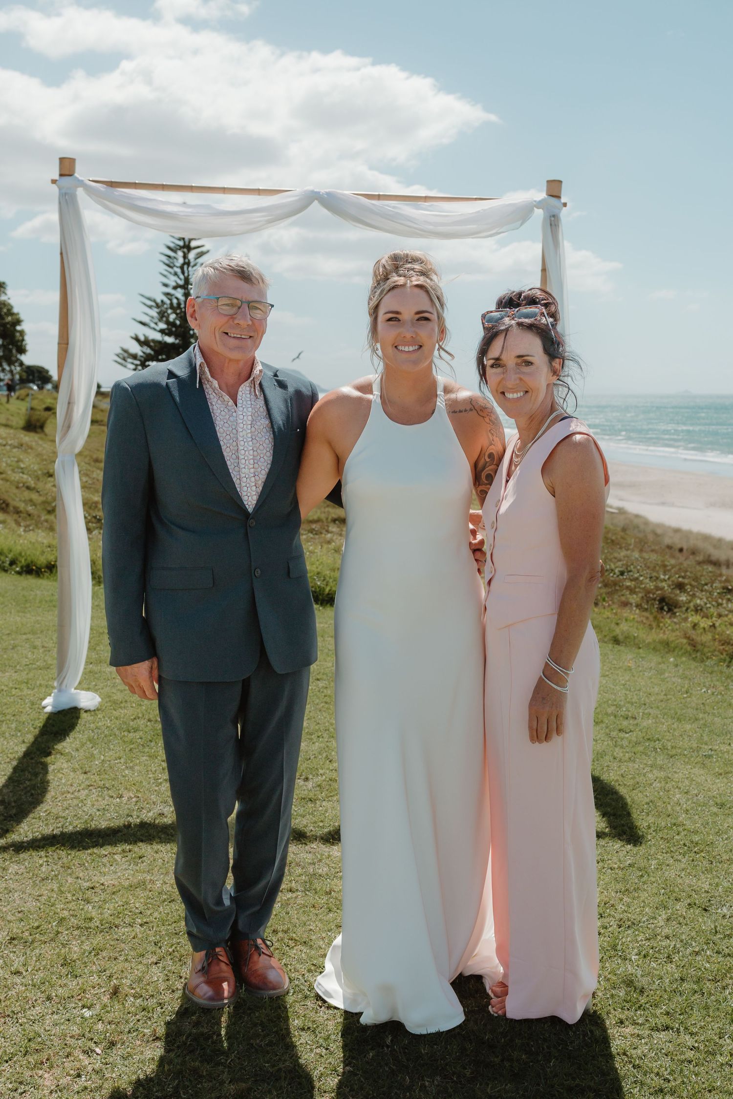 Bride in white gown poses with parents under floral arch at outdoor beach wedding ceremony.