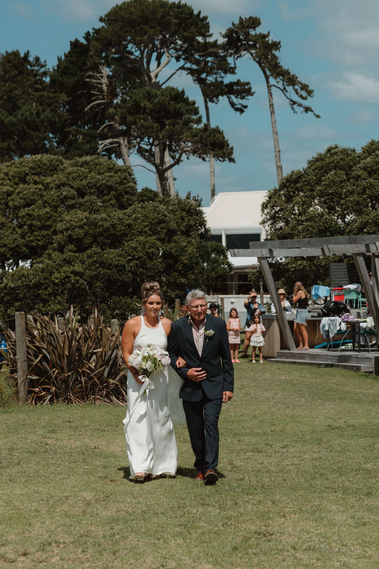 Bride in white gown walks arm-in-arm with escort down outdoor aisle at garden wedding ceremony.
