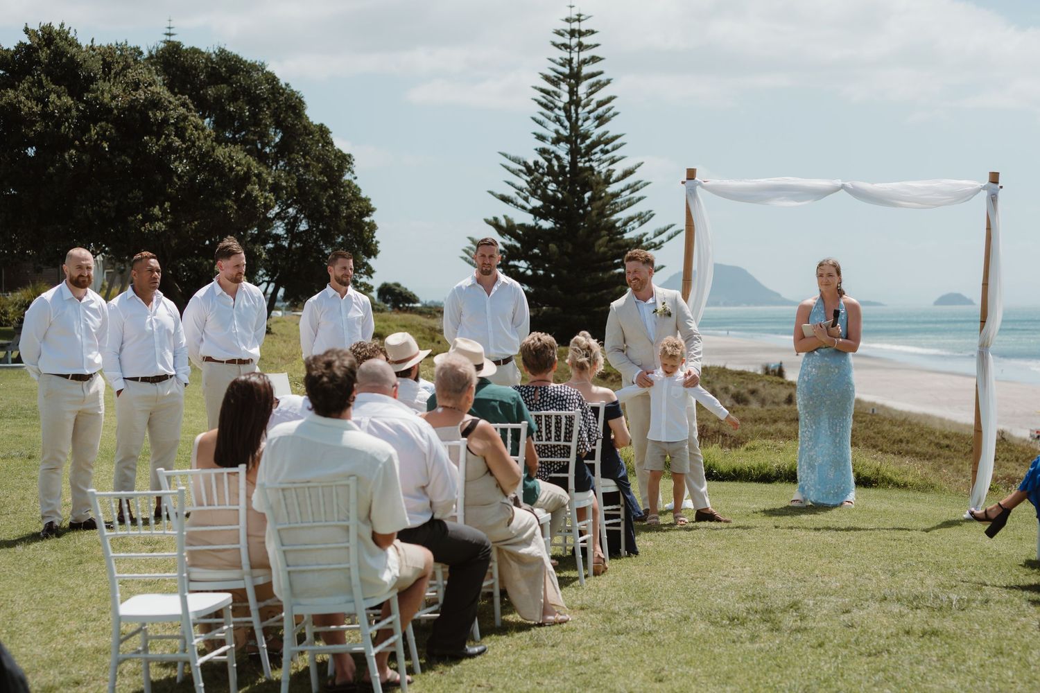 Outdoor beach wedding ceremony with groomsmen, guests seated on white chairs, and a decorated arch by the ocean.