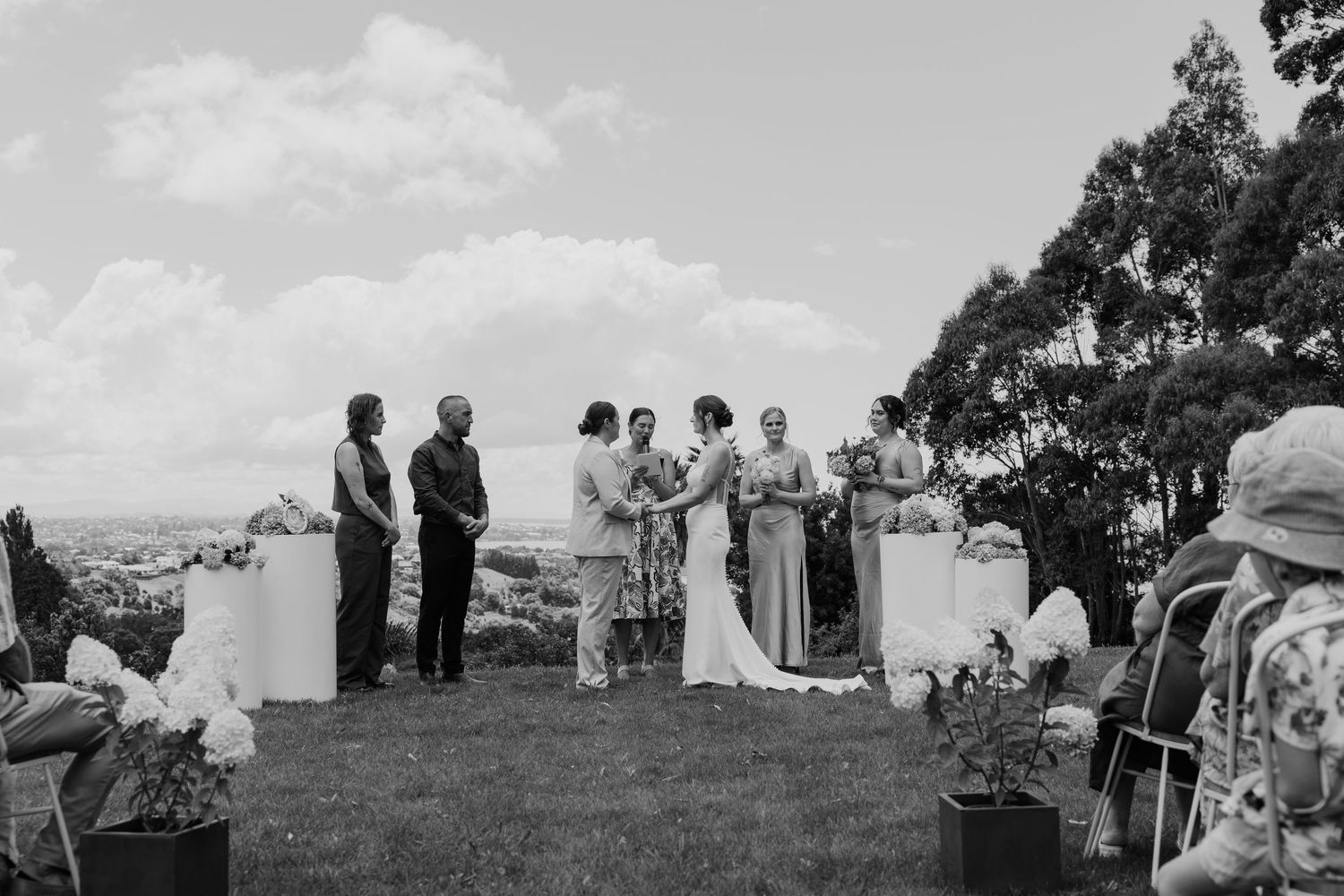 Black and white outdoor wedding ceremony on a hilltop with bridal party and guests gathered under a cloudy sky.