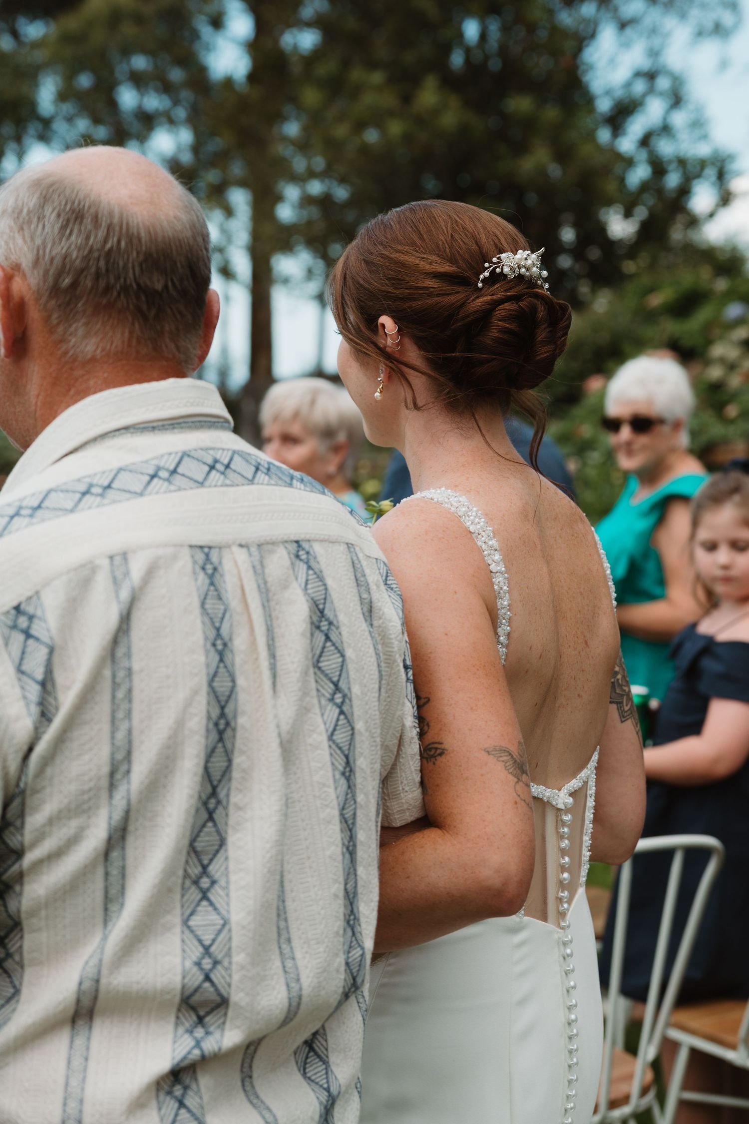 Bride in backless wedding dress with tattoo walks with older man at outdoor wedding ceremony.