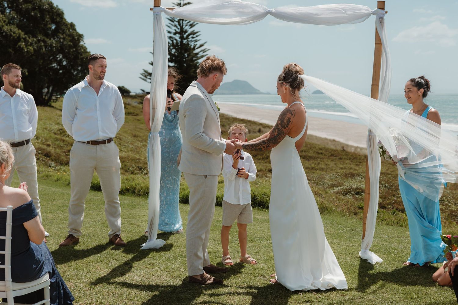 Outdoor beach wedding ceremony with couple exchanging vows under a white draped arch, guests watching on a green hillside.