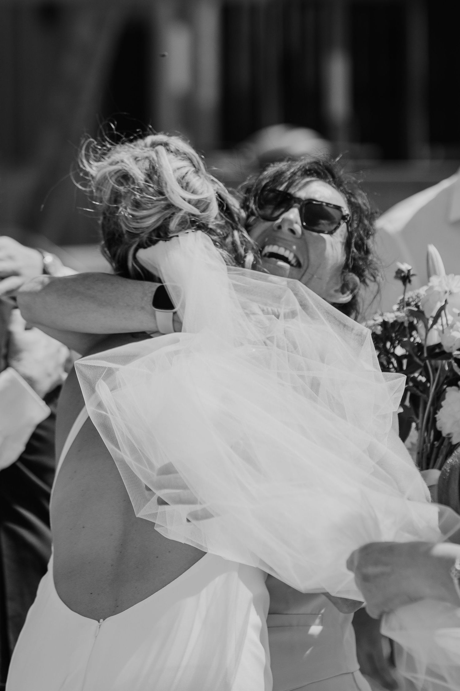 Black and white photo of a bride hugging a smiling woman in sunglasses holding a bouquet at a wedding.