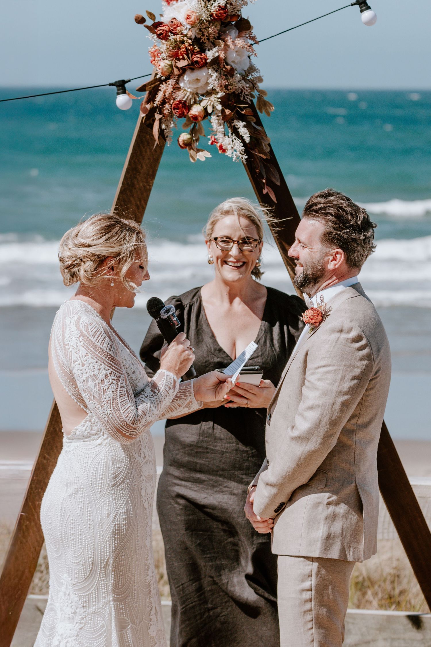 A beachside wedding ceremony takes place under a wooden arch with dried floral arrangements against turquoise ocean waves.