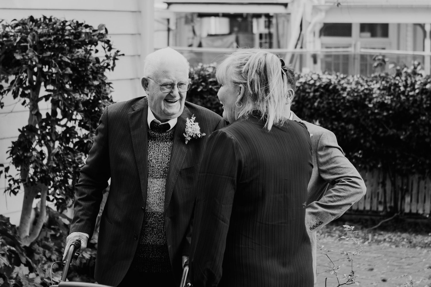 Two people share a candid black and white moment together in a residential garden setting with bushes and a fence.