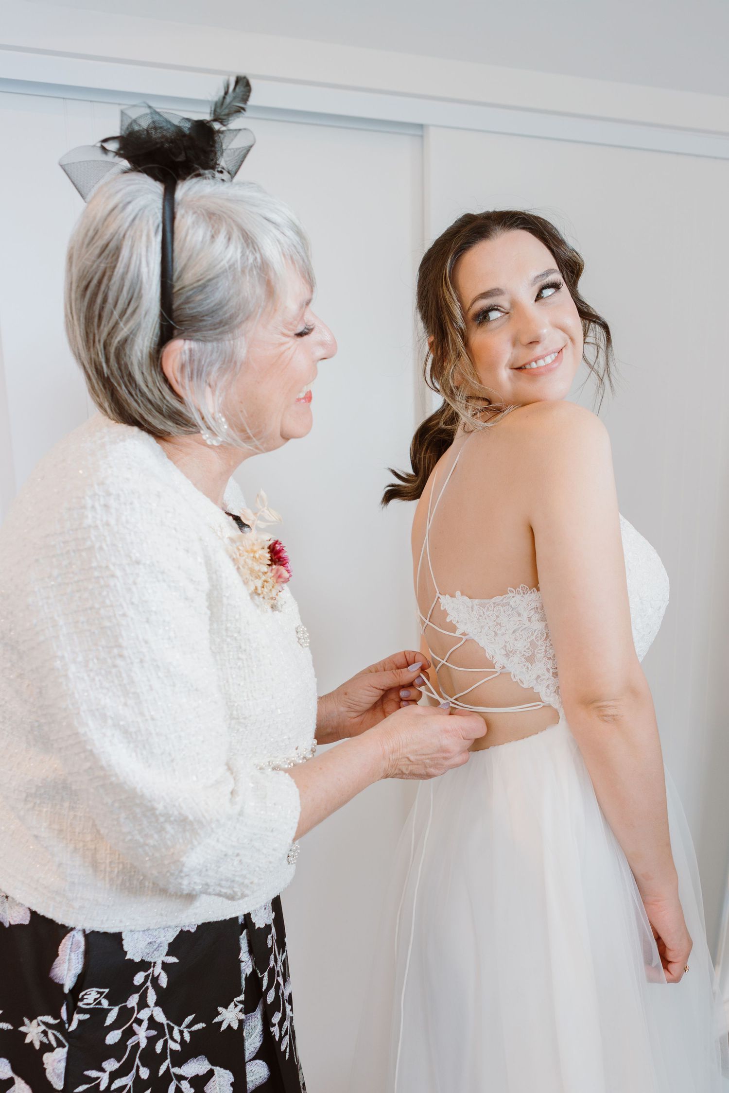 A woman in a white wedding dress smiles while getting help with the back of her gown from someone in a white cardigan.