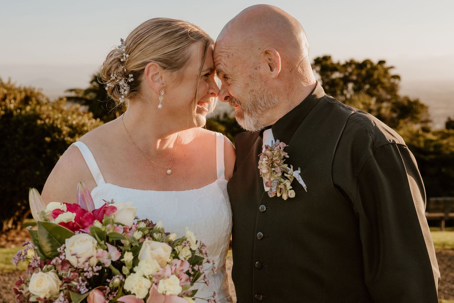 A romantic wedding couple shares an intimate moment at sunset while the bride holds a pink and white floral bouquet.