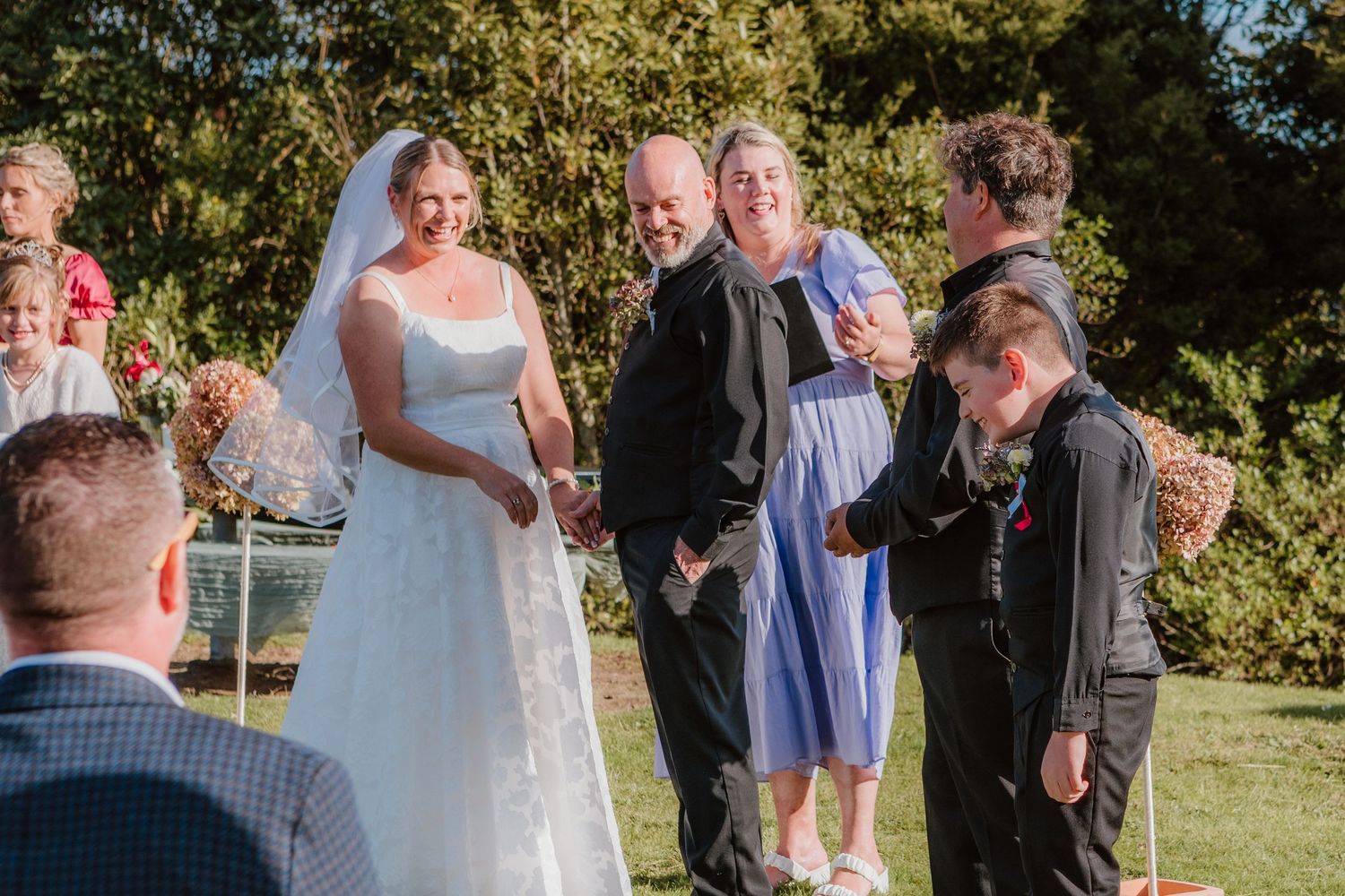 A relaxed outdoor wedding ceremony takes place in a sunny garden setting with guests gathered around on a beautiful day.