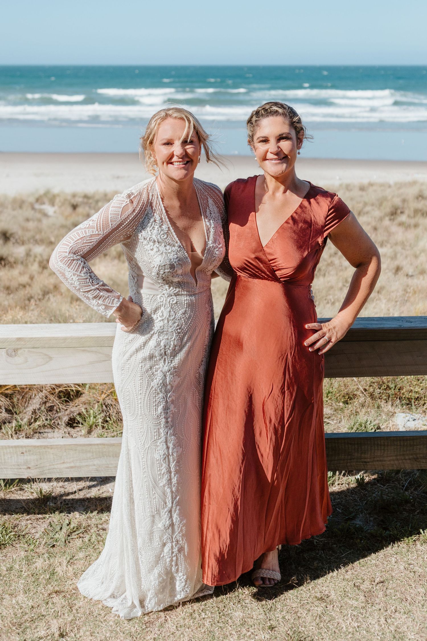 Two women pose on a beach boardwalk wearing elegant dresses in white lace and terracotta colors. Papamoa Surf Lifesaving Club Wedding