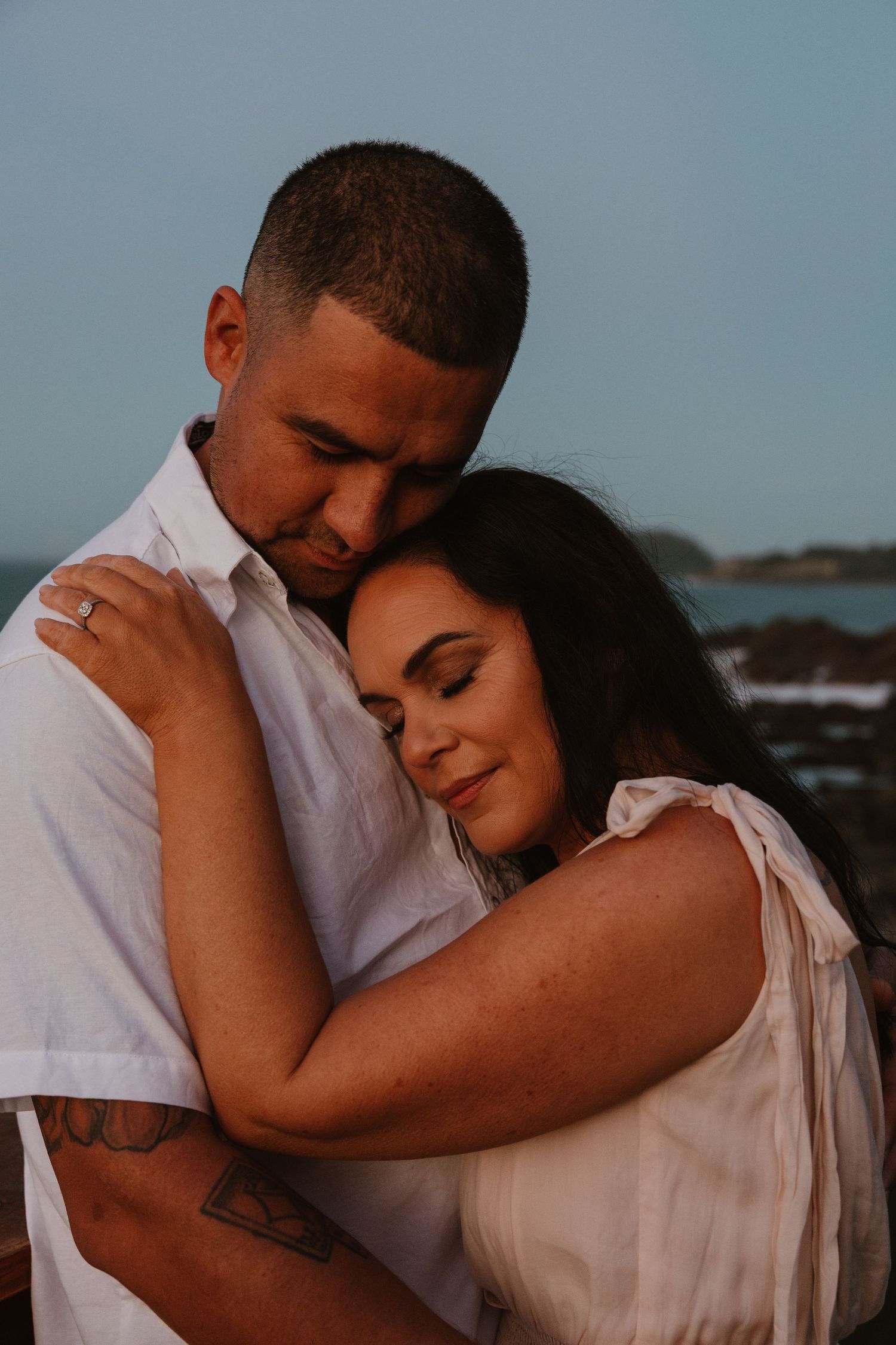 A couple in white outfits shares an intimate embrace against a coastal backdrop during sunset.