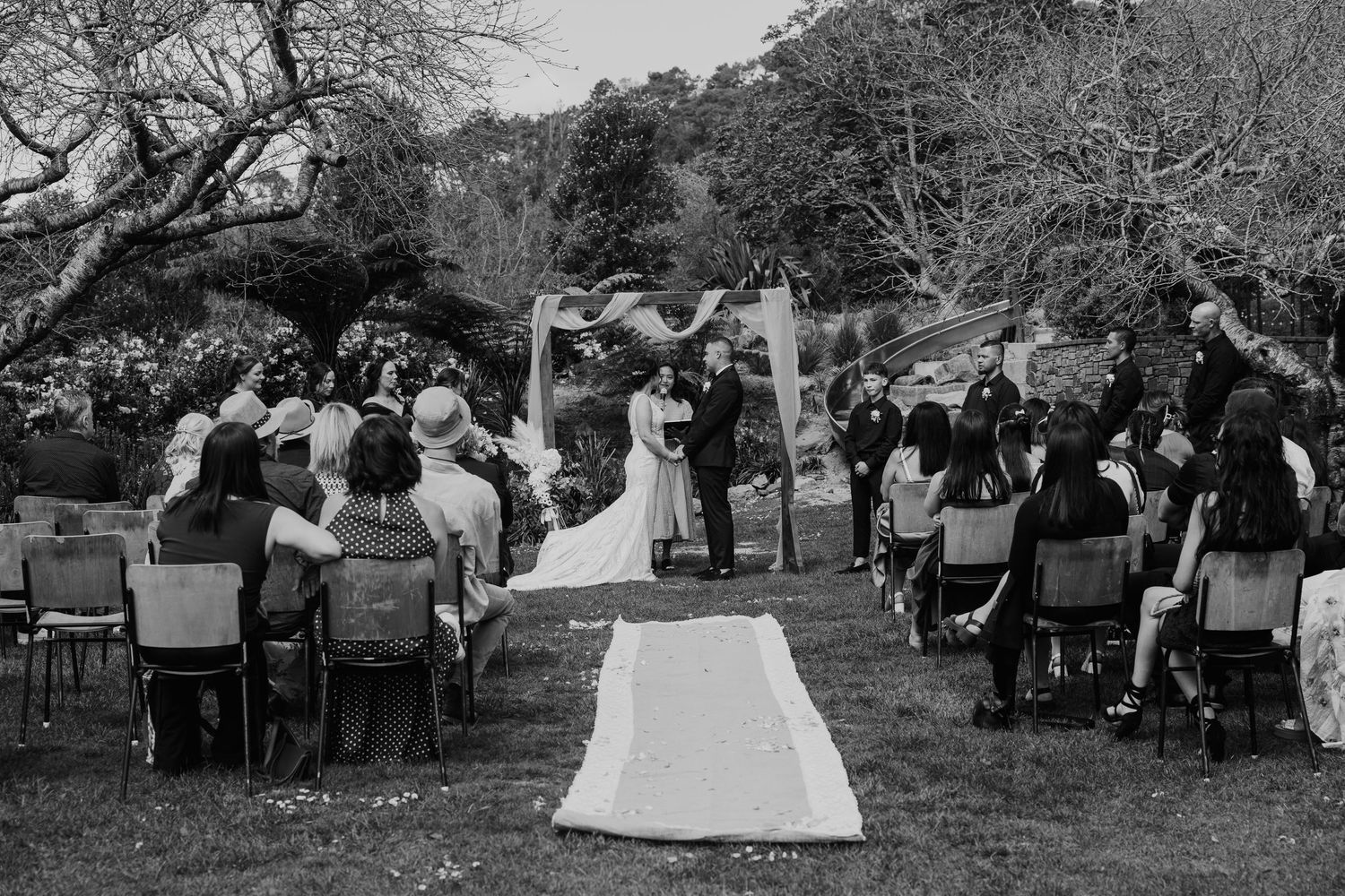 A black and white wedding ceremony takes place outdoors with guests seated in rows under trees.
