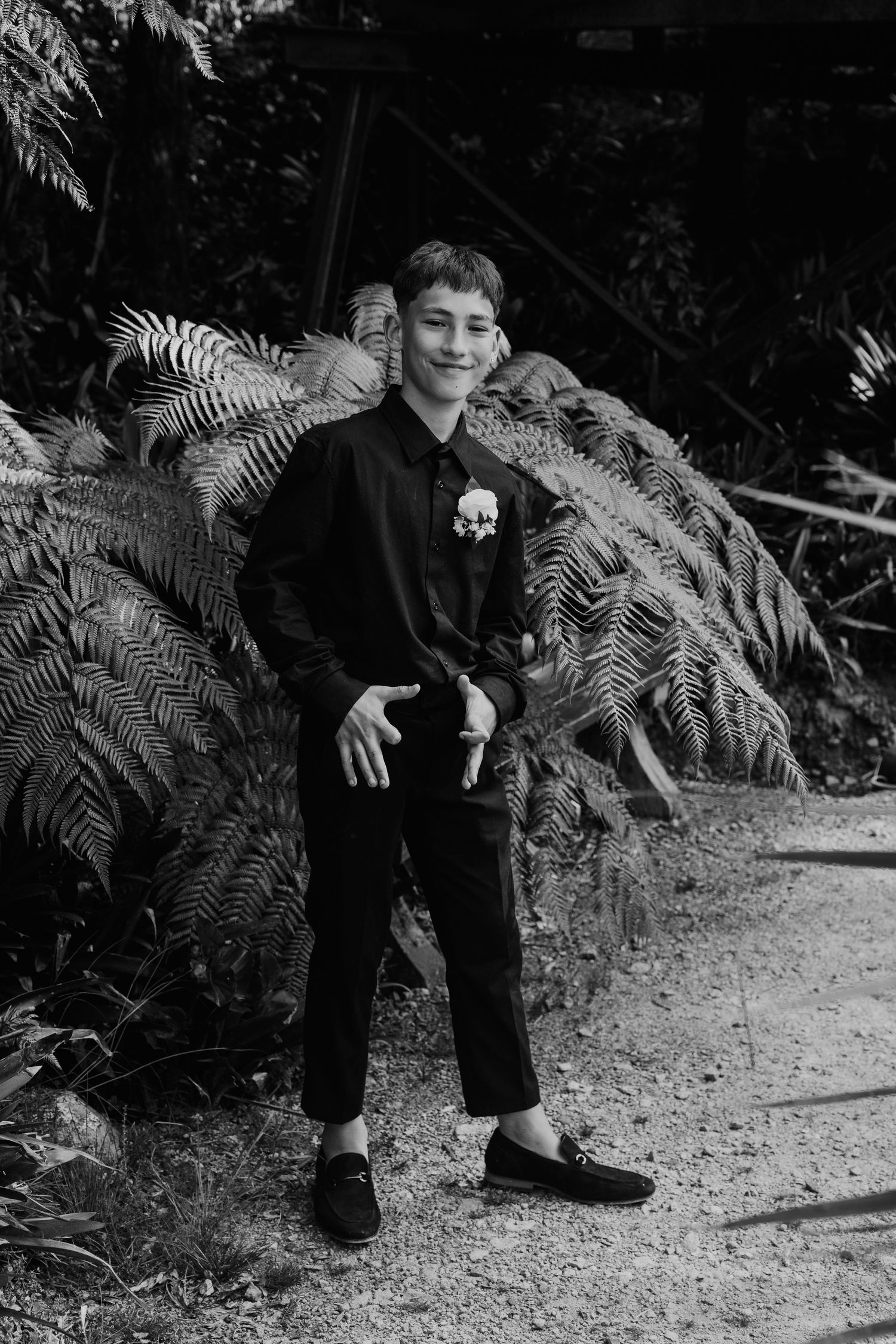 A young person in dark clothing crouches by ferns on a gravel path in a black and white nature scene.