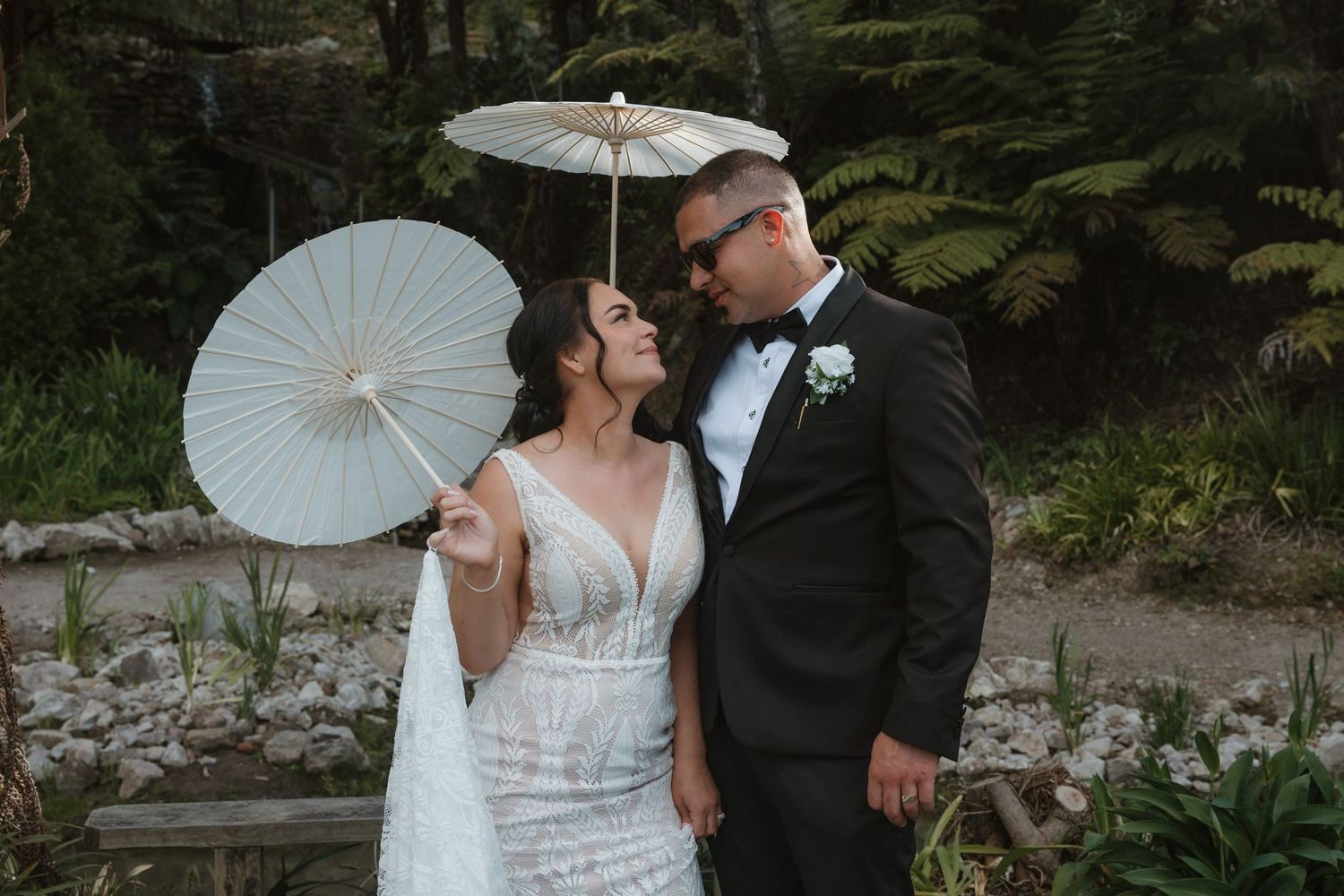 A romantic couple shares an intimate moment under white parasols in a lush garden setting during their wedding celebration.