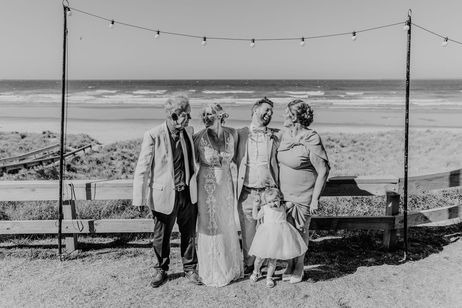 Black and white beach wedding photo of small group standing beneath string lights along coastal shore.