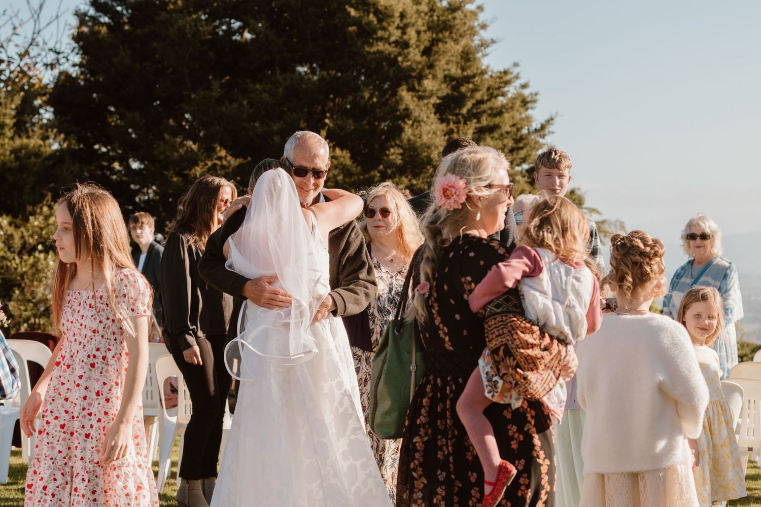 Wedding guests outdoors at a sunset celebration with attendees in formal attire gathered on a sunny afternoon.
