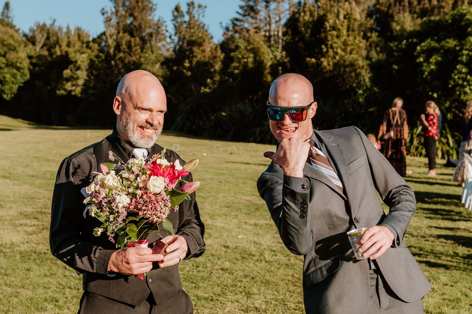Two men in dark suits stand in a grassy field, one holding a floral bouquet while the other adjusts his sunglasses.
