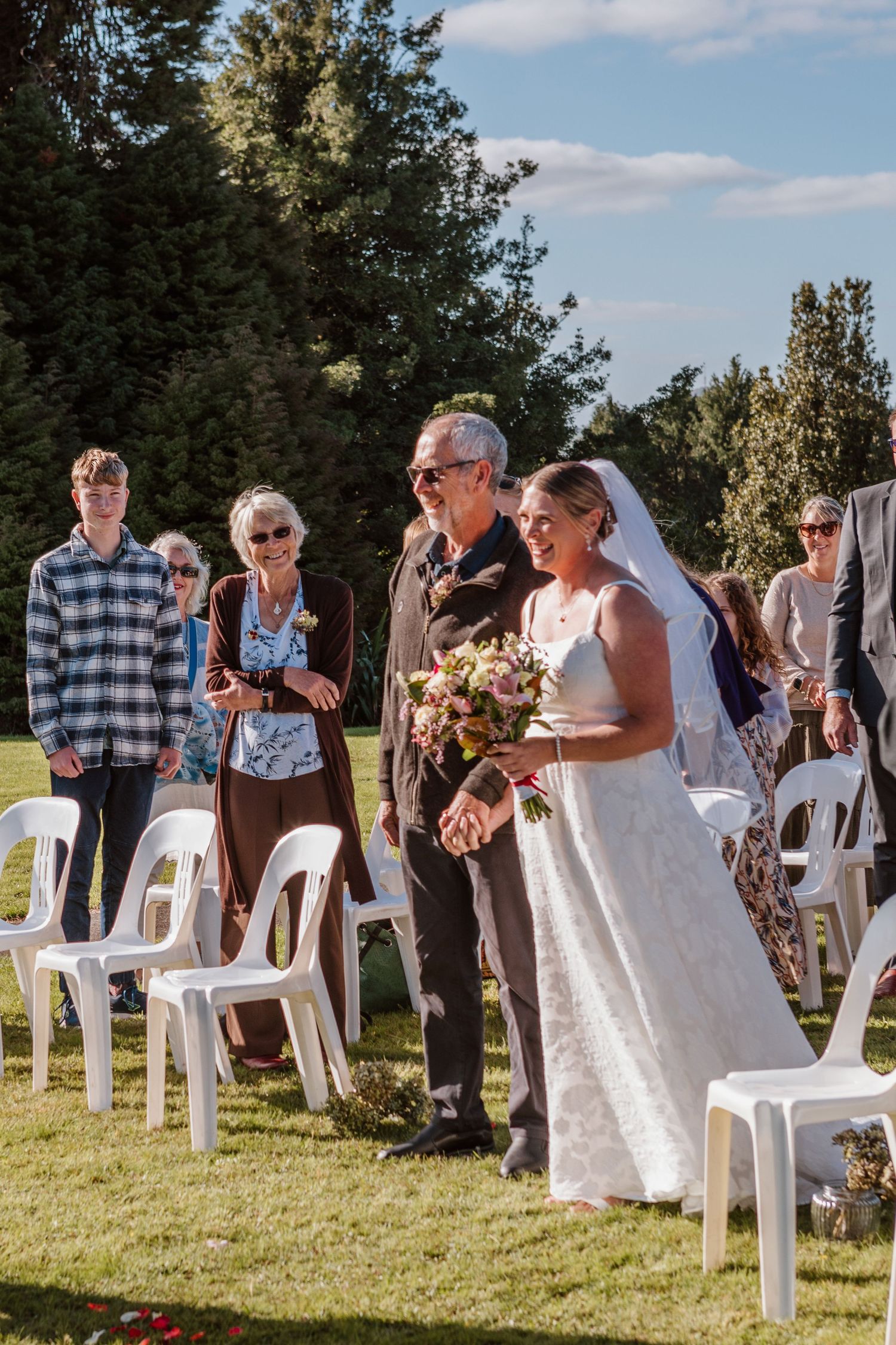 Outdoor wedding ceremony on a sunny day with white chairs set up on a lawn surrounded by trees.