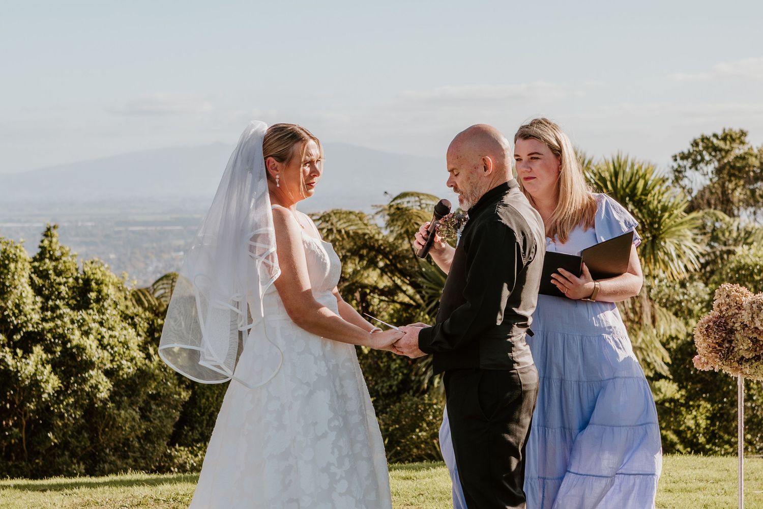 An outdoor wedding ceremony takes place on a hilltop with mountains and lush greenery in the background.