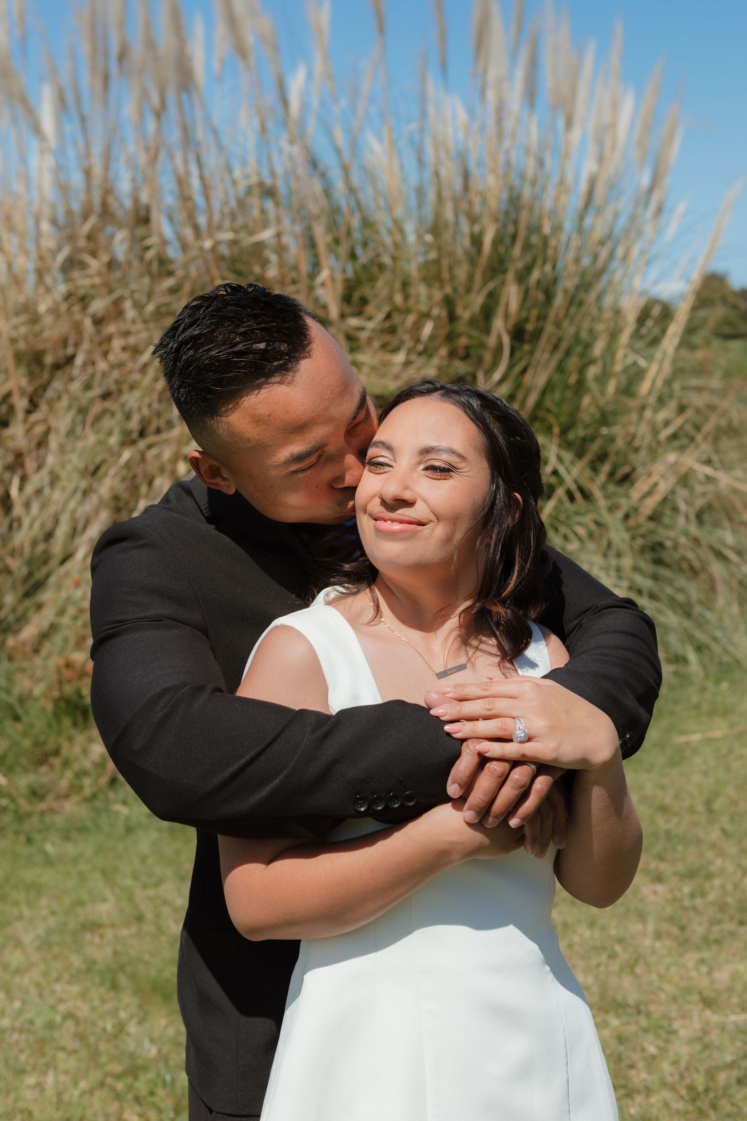 A romantic couple embraces in front of tall grass on a sunny day, showing tender affection in a series of candid poses.