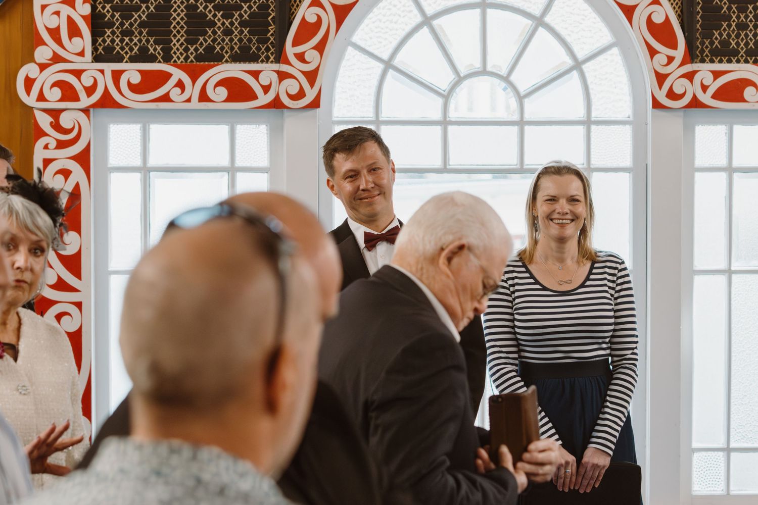 People gather near decorative windows with ornate red trim and arched design during an indoor event.
