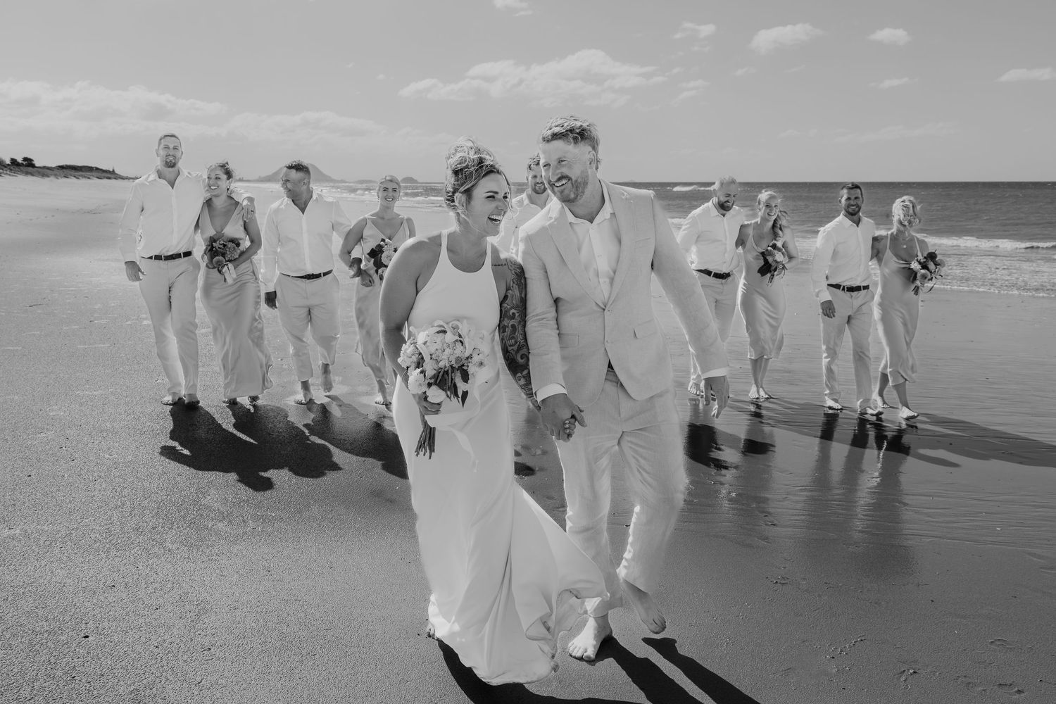 Black and white beach wedding photo of bride and groom leading wedding party along sandy shoreline.