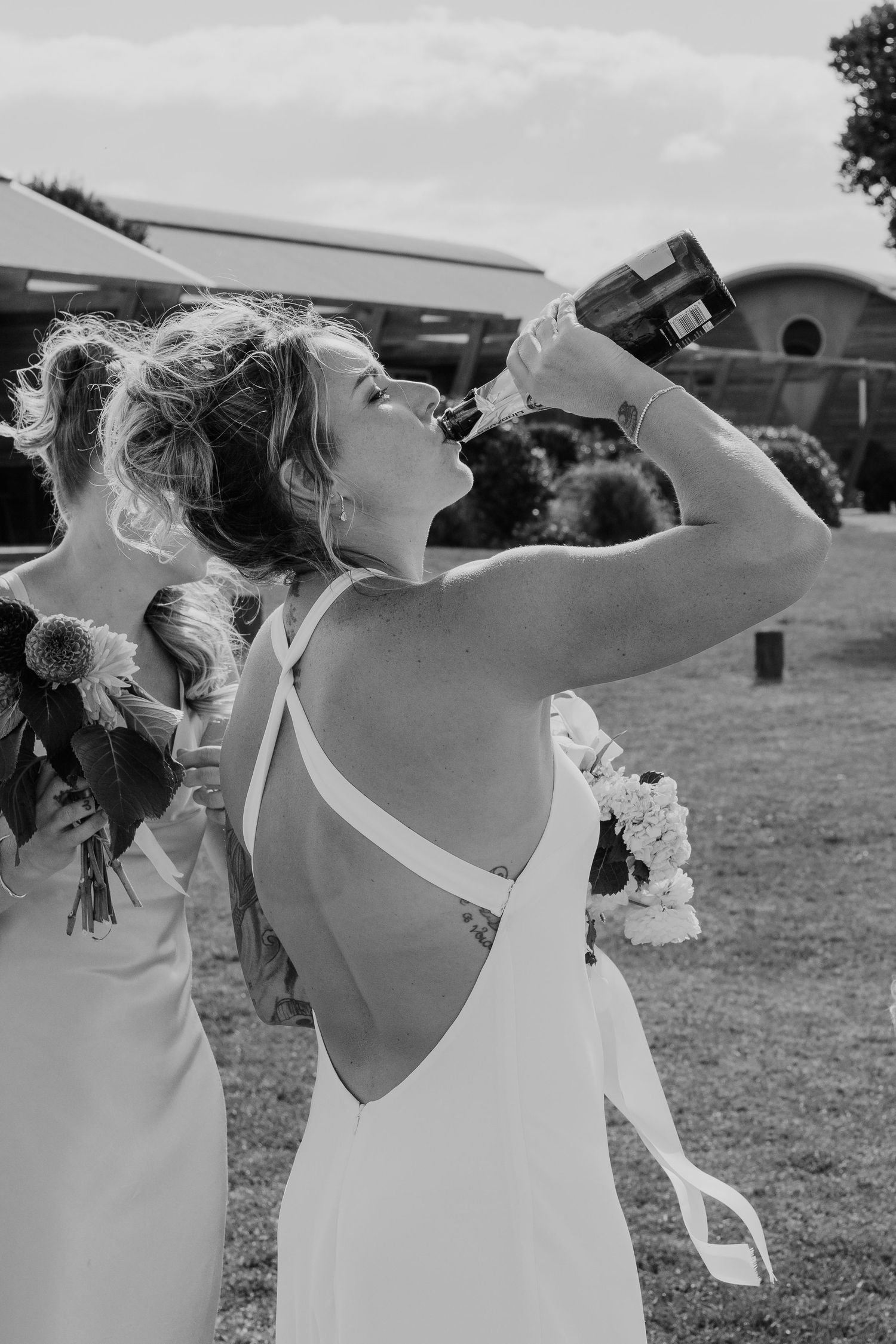 Bride in backless wedding dress drinks champagne from bottle outdoors at wedding celebration in black and white photo.