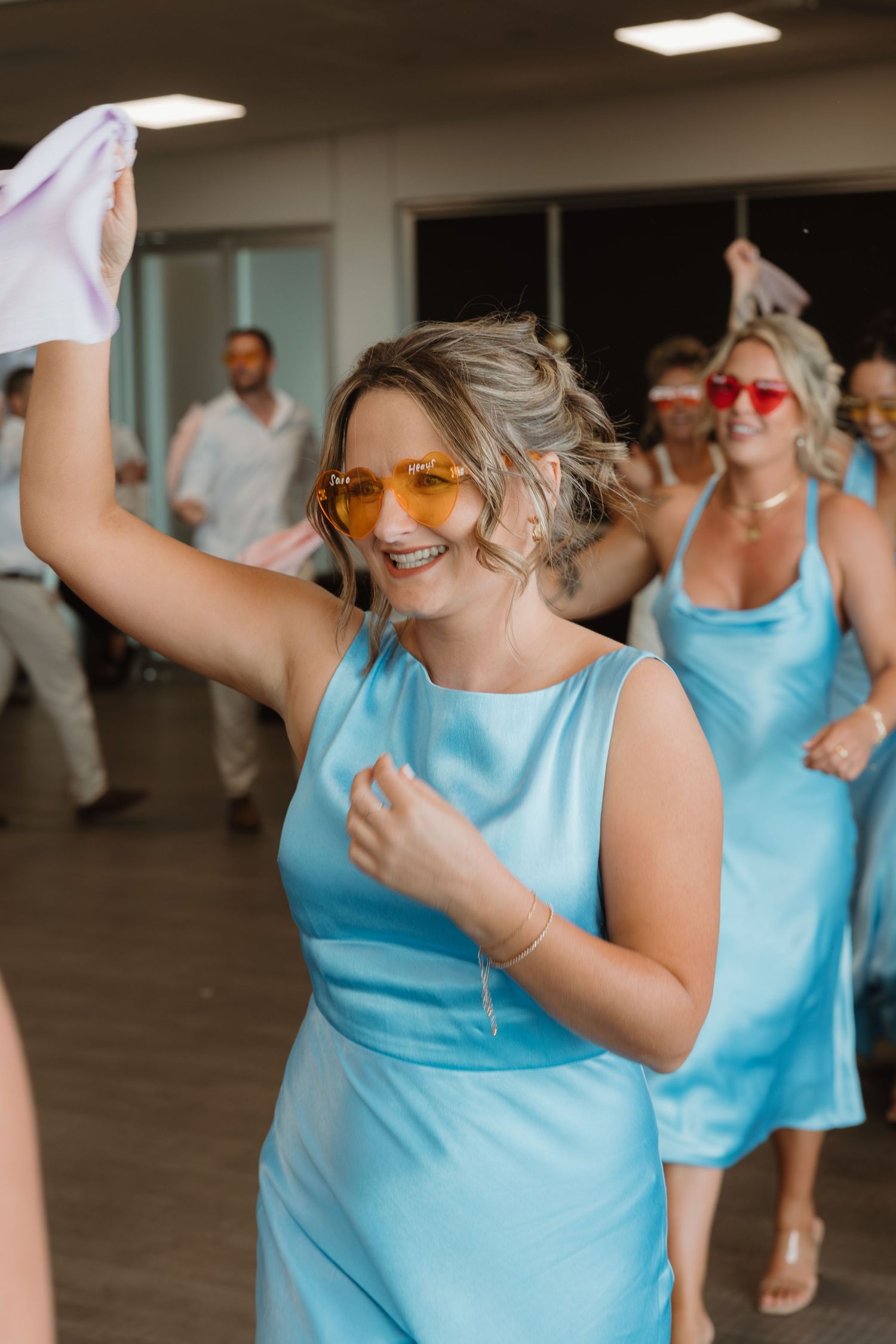 Bridesmaids in light blue satin dresses dancing joyfully at a wedding reception, wearing colorful sunglasses.