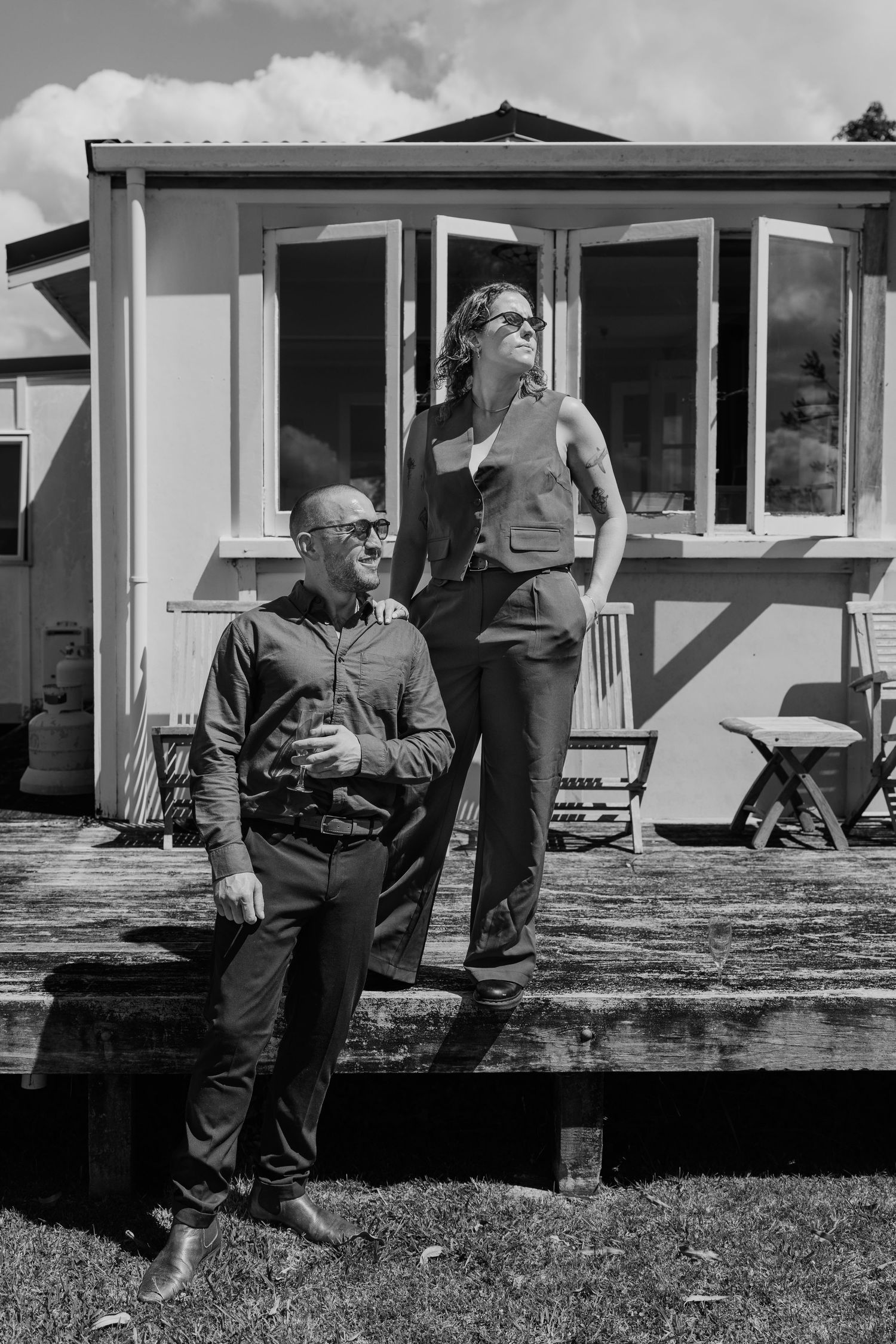 Black and white photo of a man and woman posing outdoors on a deck in front of a small building on a sunny day.