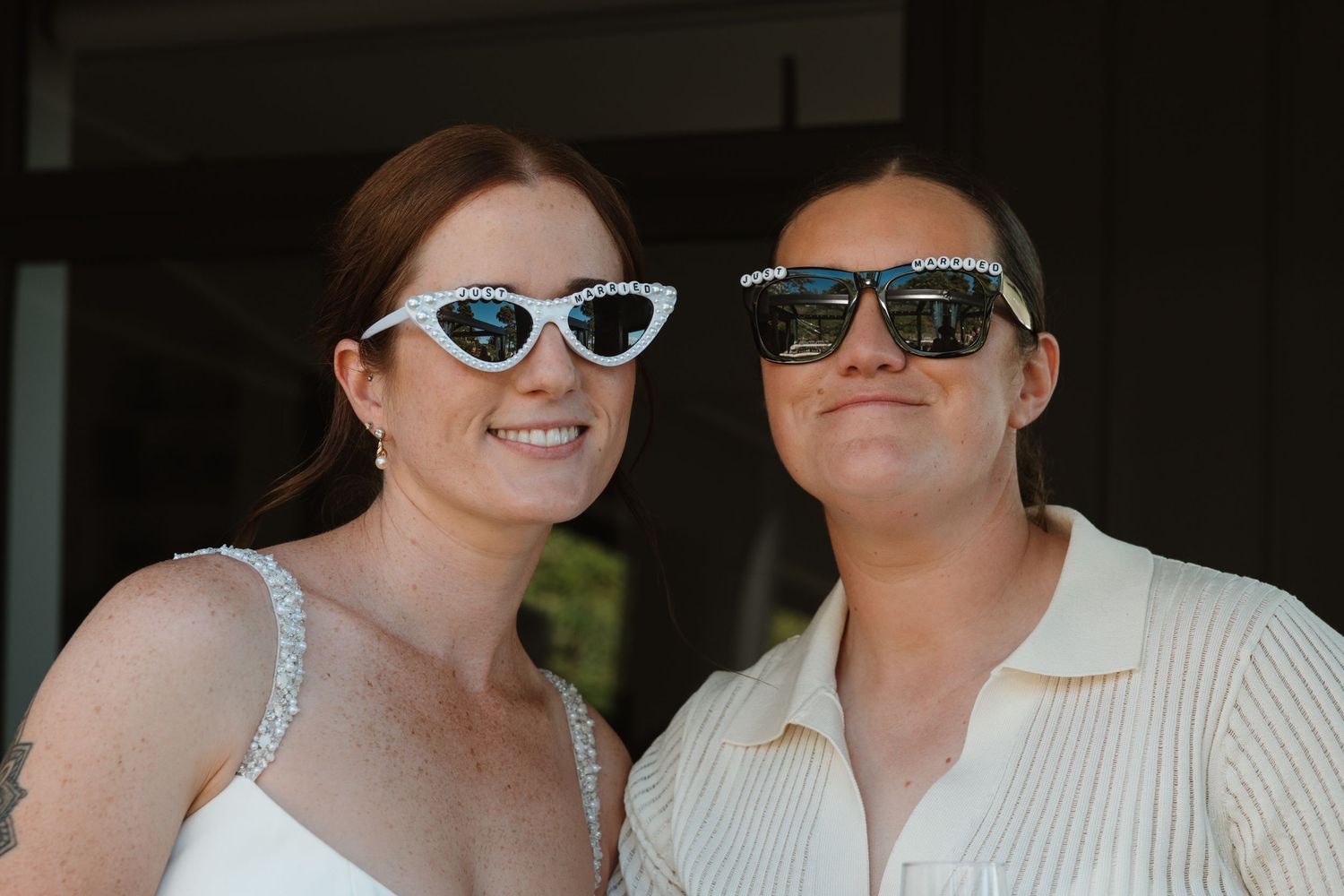 Two people smiling and wearing decorative sunglasses, one in a white dress, posing together outdoors.