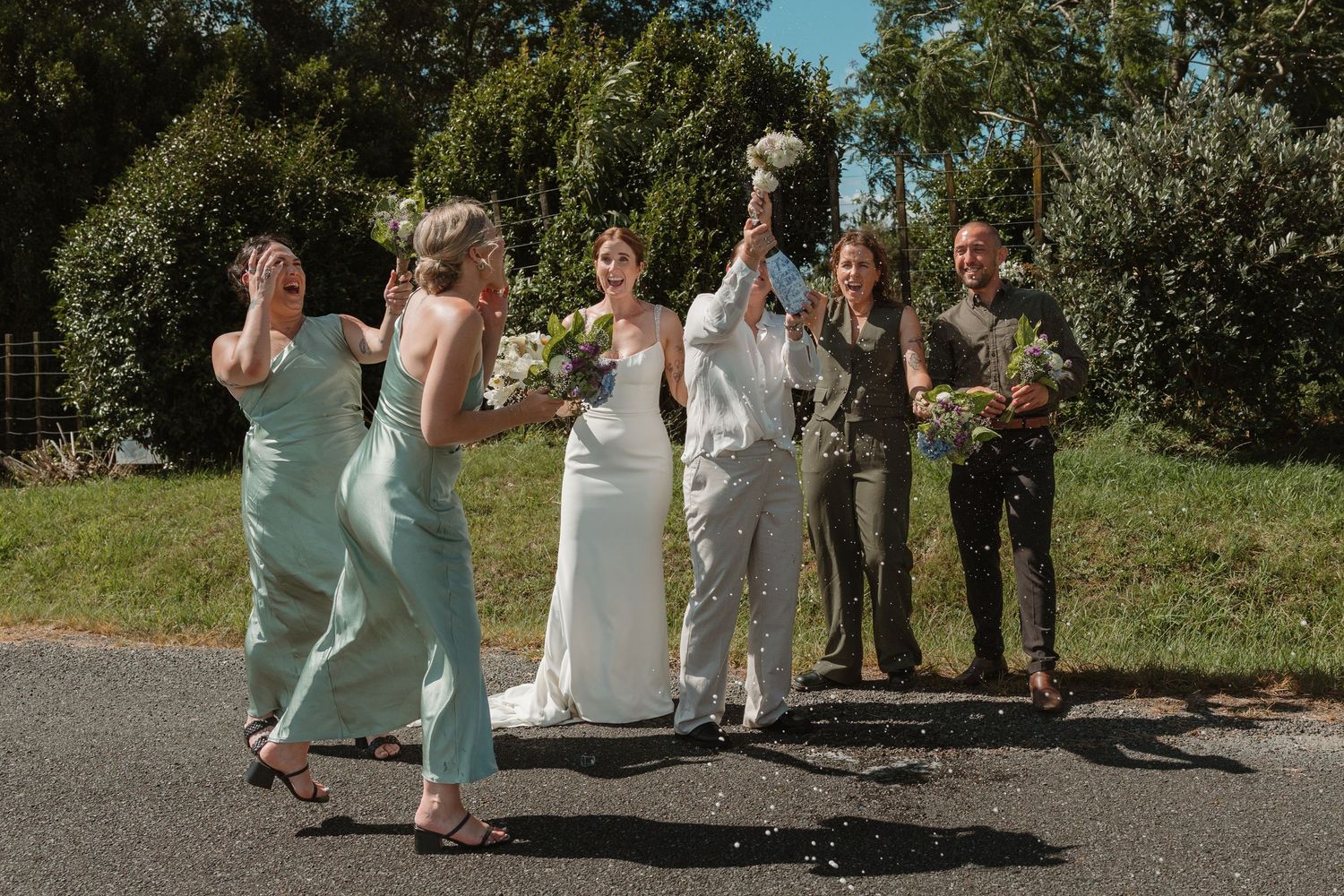 Wedding party celebrating outdoors, brides in white suits with bridesmaids in mint green and groomsmen on a sunny day.