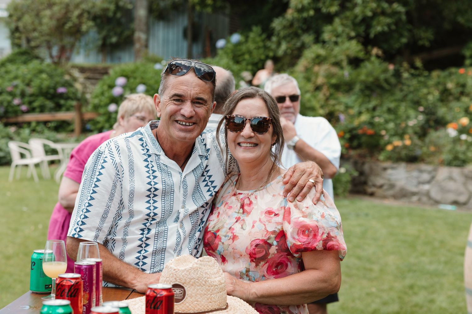 Smiling couple posing together at an outdoor garden party, woman in floral dress and man in striped shirt.