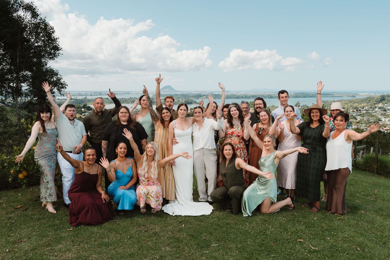 Wedding group photo with guests cheering outdoors, scenic ocean and mountain views in background on a sunny day.