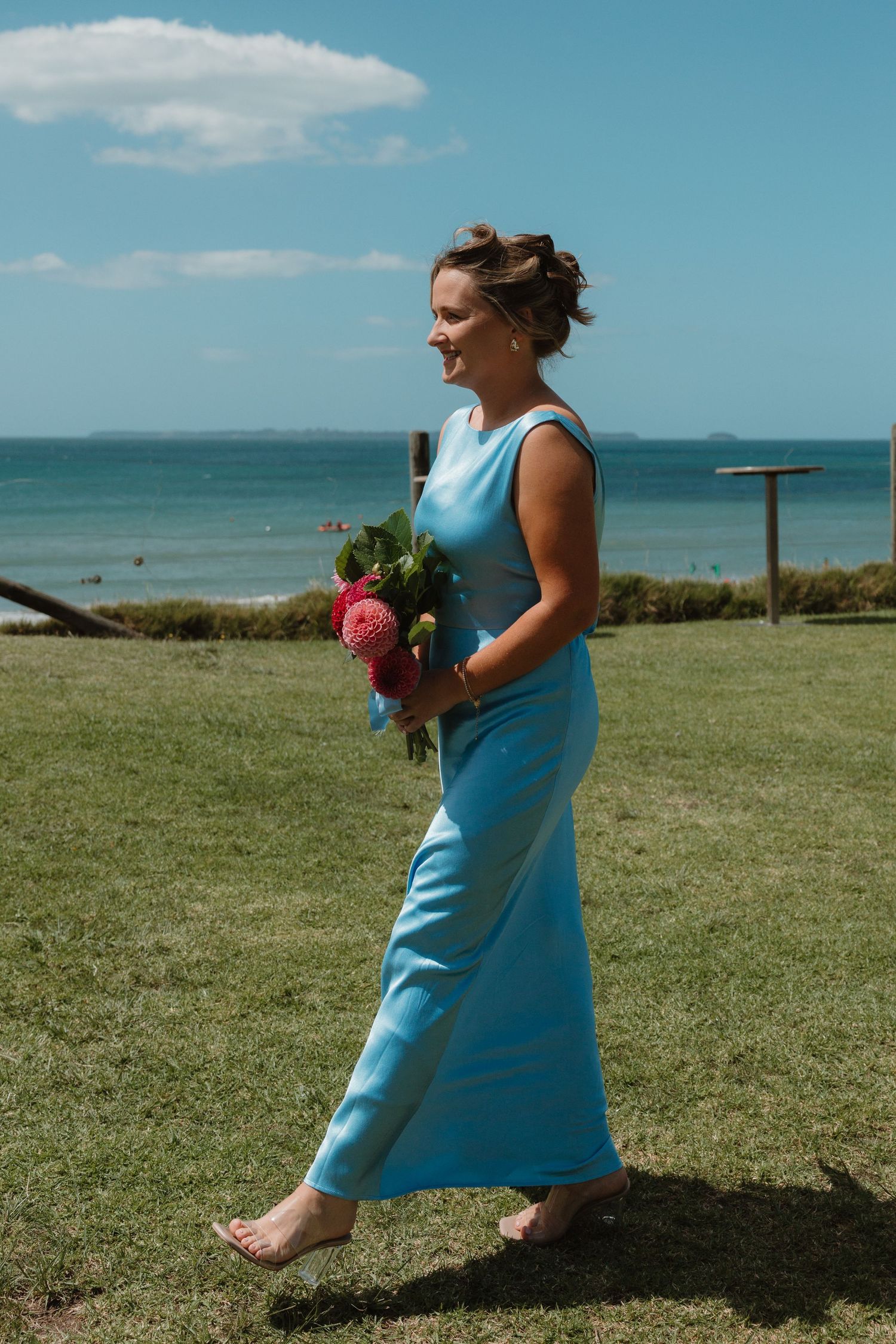 Smiling bridesmaid in blue dress holding pink bouquet walks barefoot on green grass with ocean backdrop.