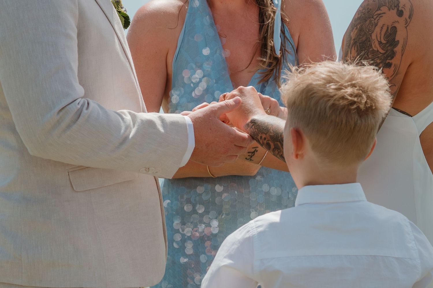 A young boy watches as a couple exchanges rings during an outdoor wedding ceremony on a sunny day.