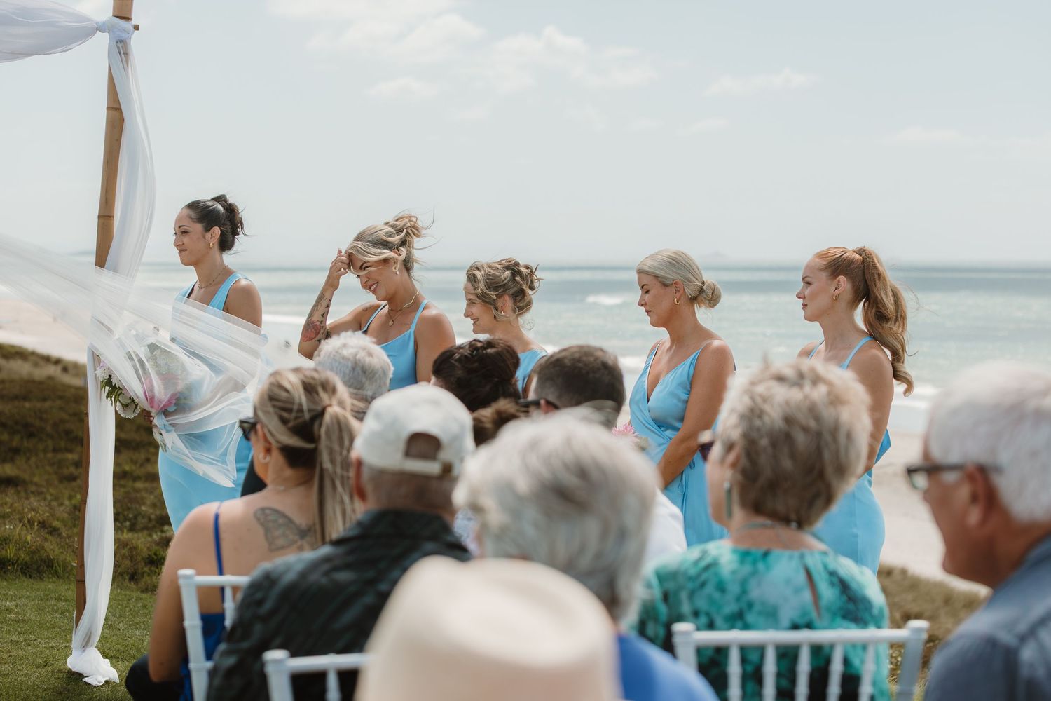 Bridesmaids in blue dresses stand at an outdoor beachside wedding ceremony with guests seated in white chairs.