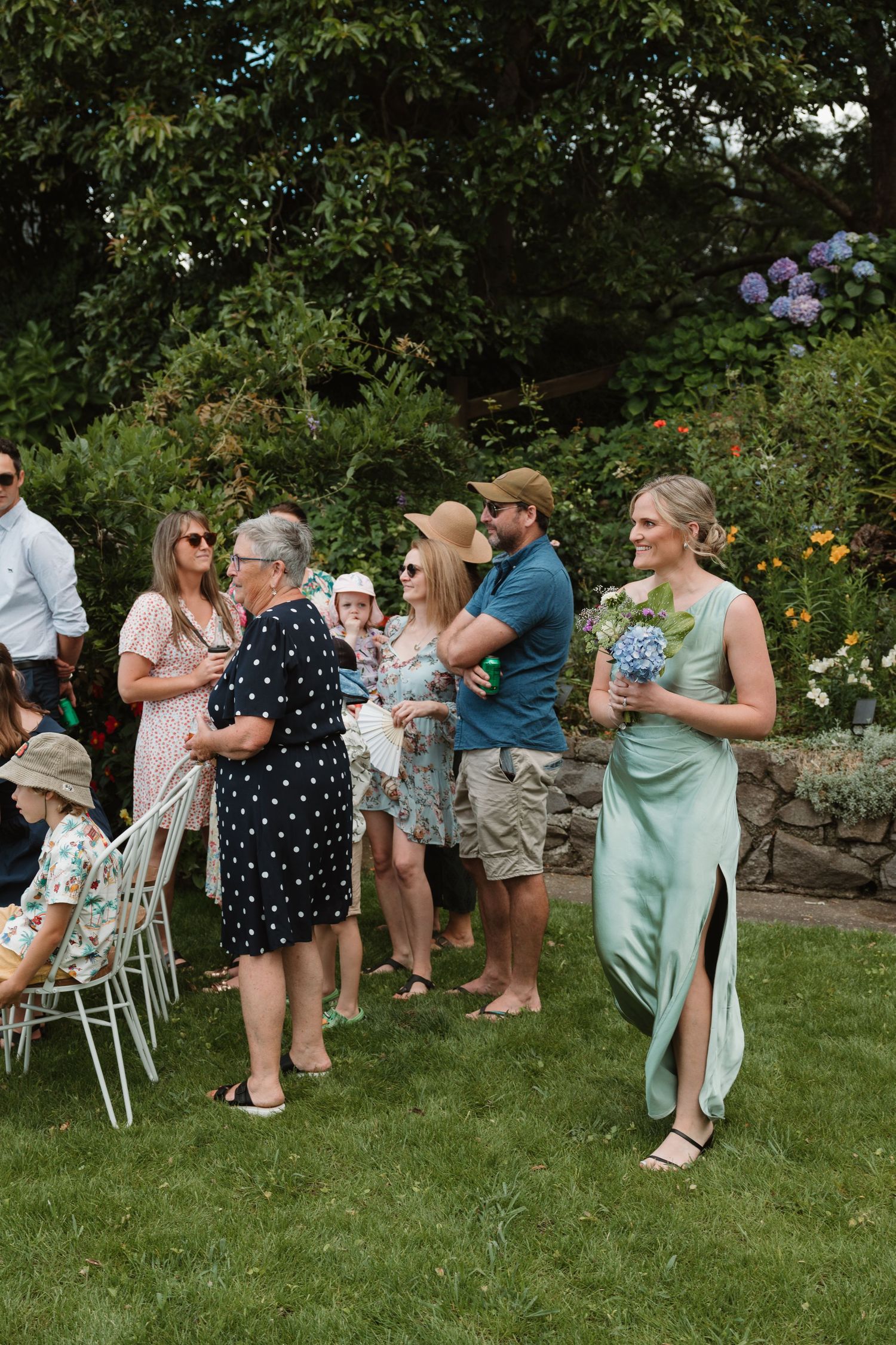 Bridesmaid in mint green dress holds bouquet at outdoor garden wedding ceremony with guests mingling on lawn.