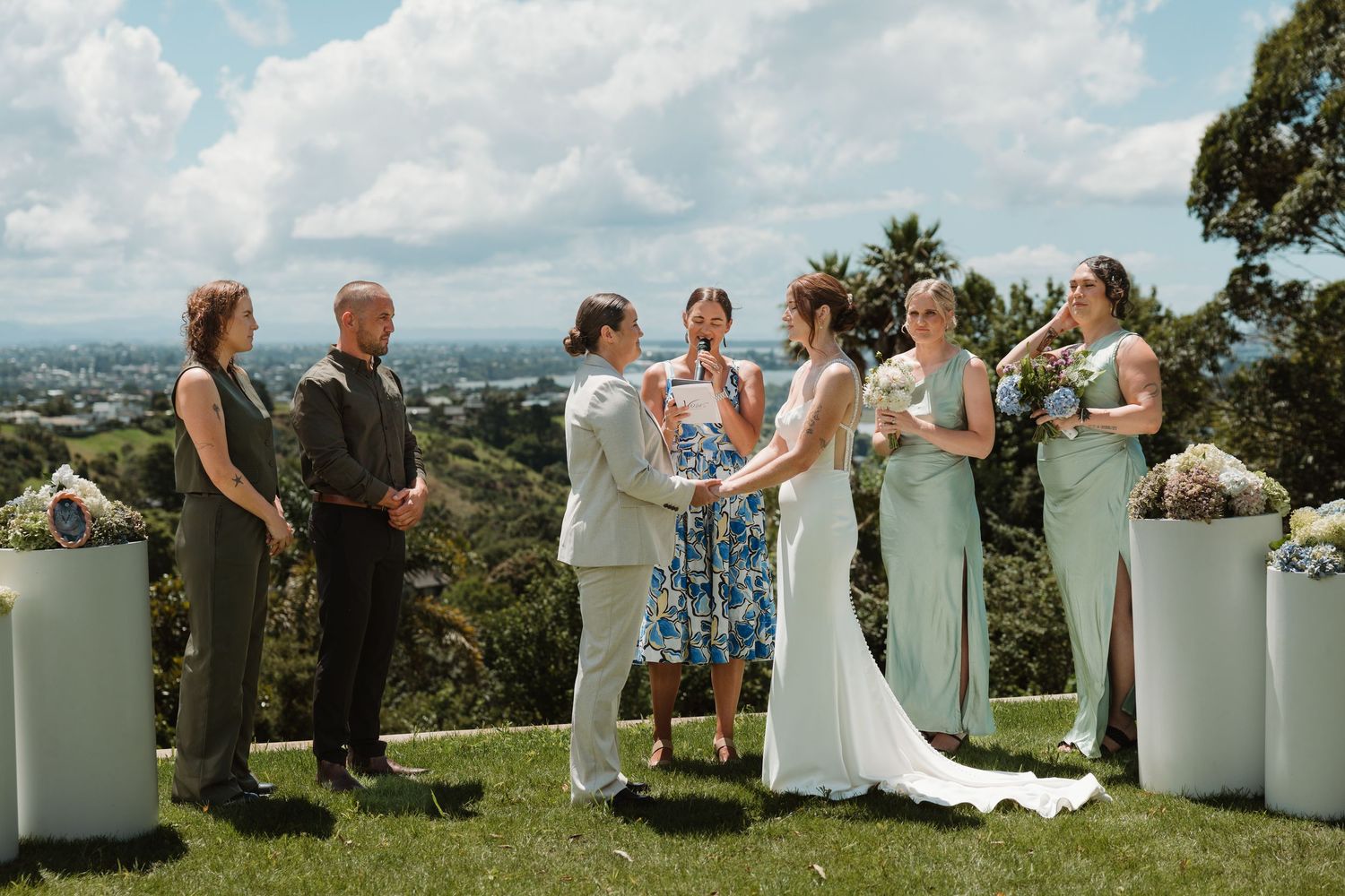 Outdoor wedding ceremony on a hilltop with bride, groom, officiant, and wedding party against a scenic city view.