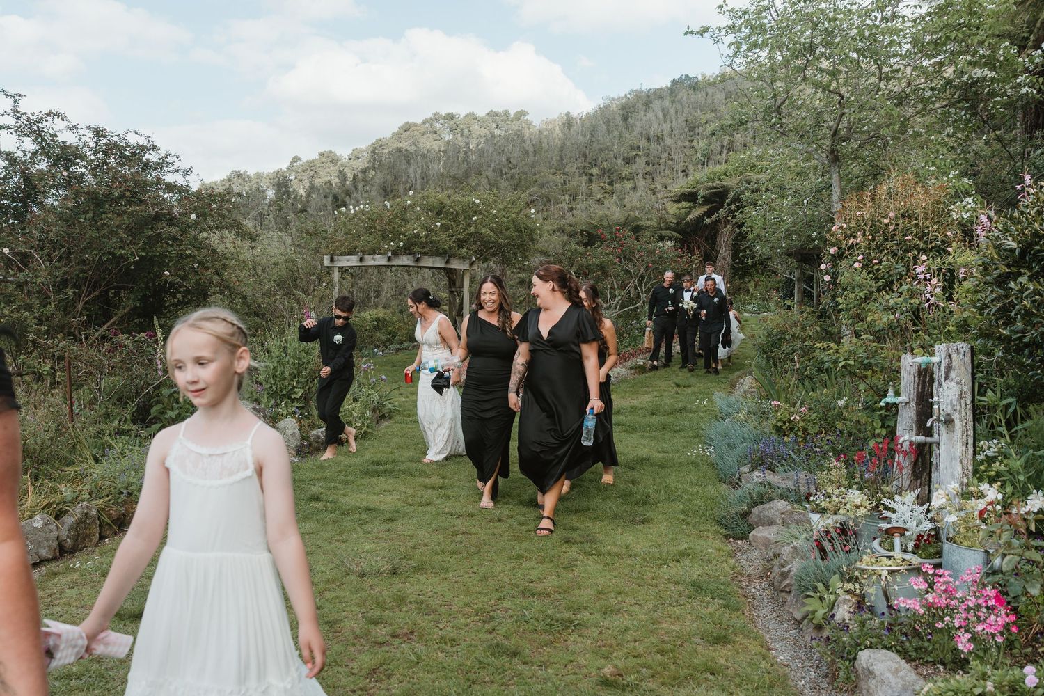 A wedding ceremony takes place in a lush green garden setting with guests gathered along a winding path.