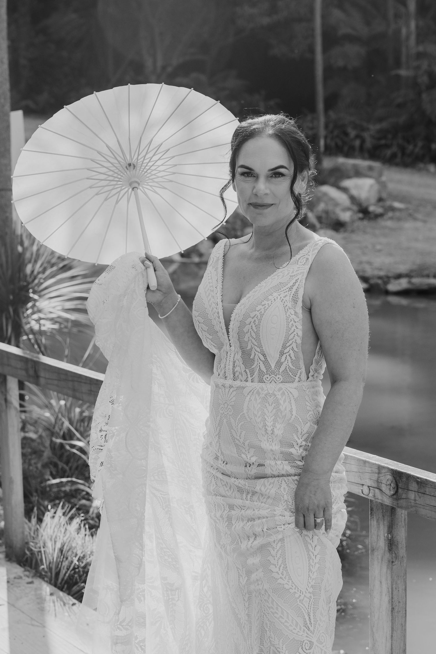 Black and white portrait of a bride in a lace wedding gown holding a white parasol in an outdoor garden setting.