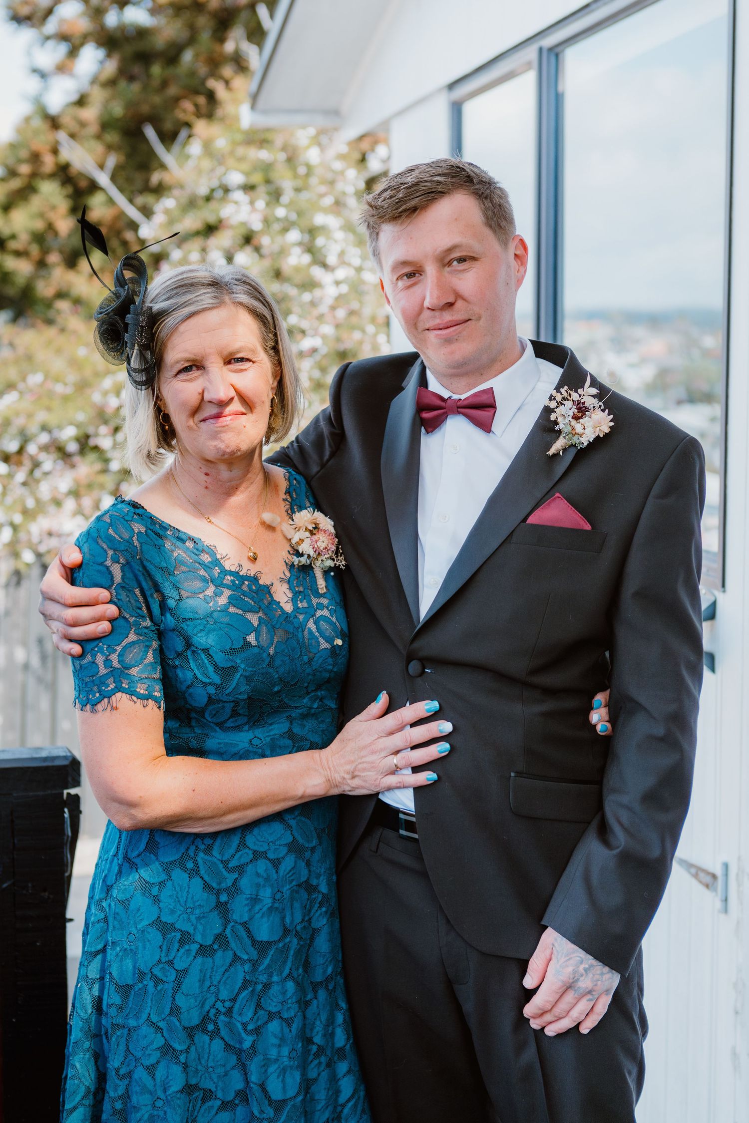 A wedding photo on a deck showing two people in formal attire, with teal lace dress and black tuxedo with burgundy bow tie.
