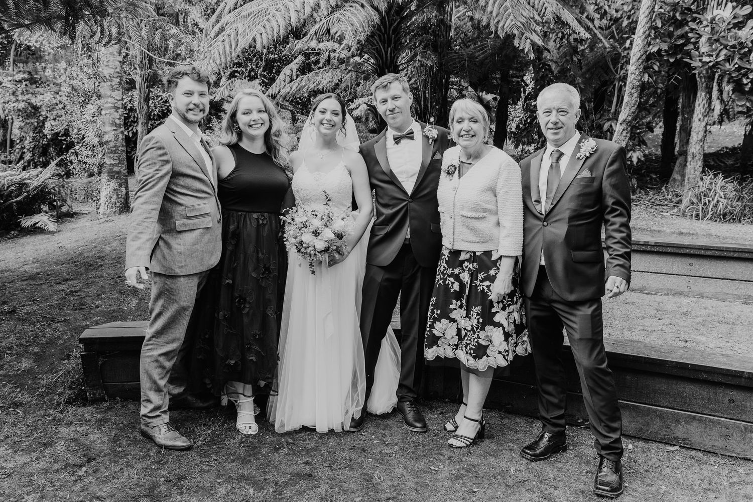 Wedding group photo with six people gathered outdoors under palm trees.