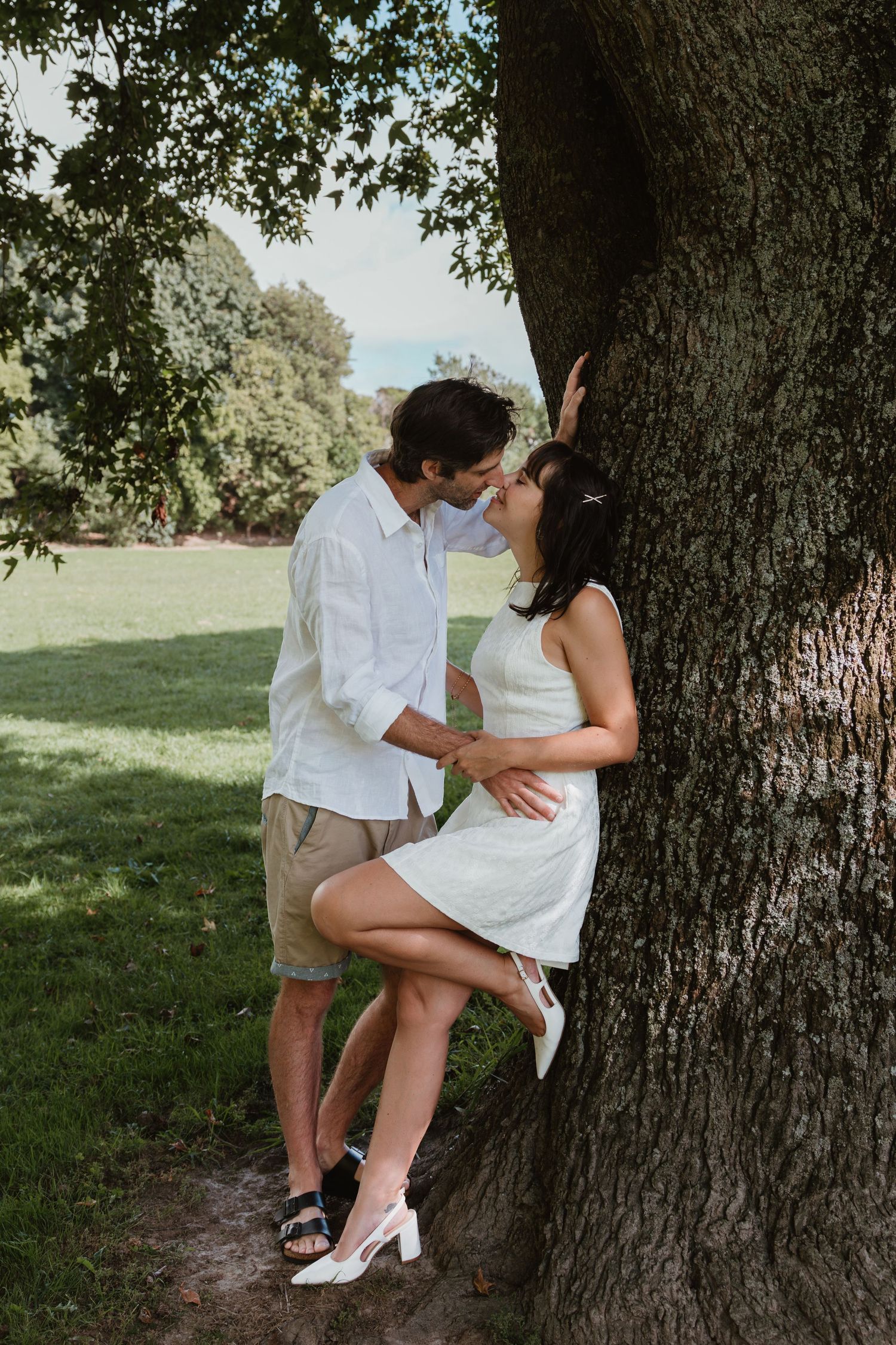 Couple sharing a romantic kiss leaning against a large tree in a lush green park on a sunny day.