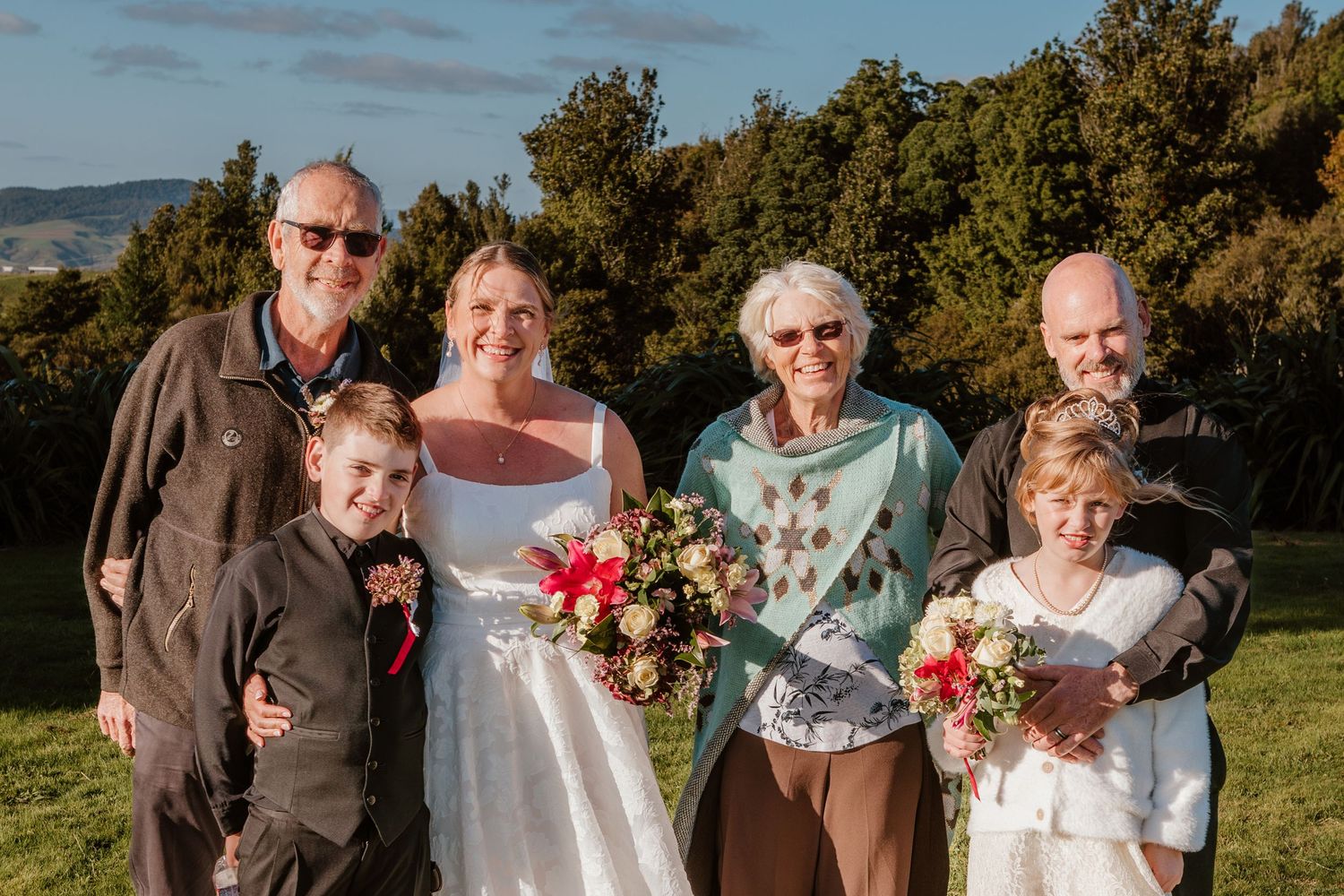 Wedding portrait of a group with bride holding bouquet outdoors against scenic countryside backdrop on sunny day.