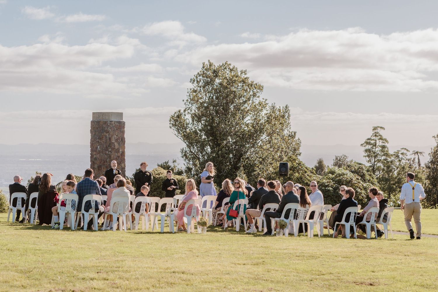 Outdoor wedding ceremony setup on a grassy hilltop with rows of white chairs and guests gathered under cloudy skies.