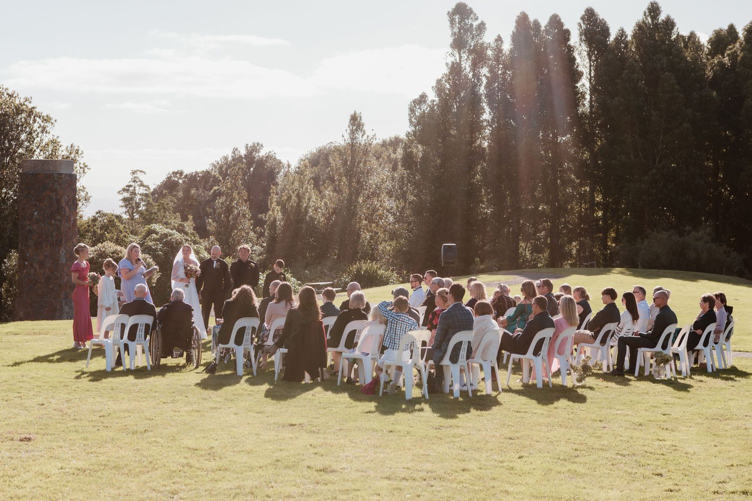 Group of wedding guests gather together for photos in a sunny outdoor field with tall trees in the background.