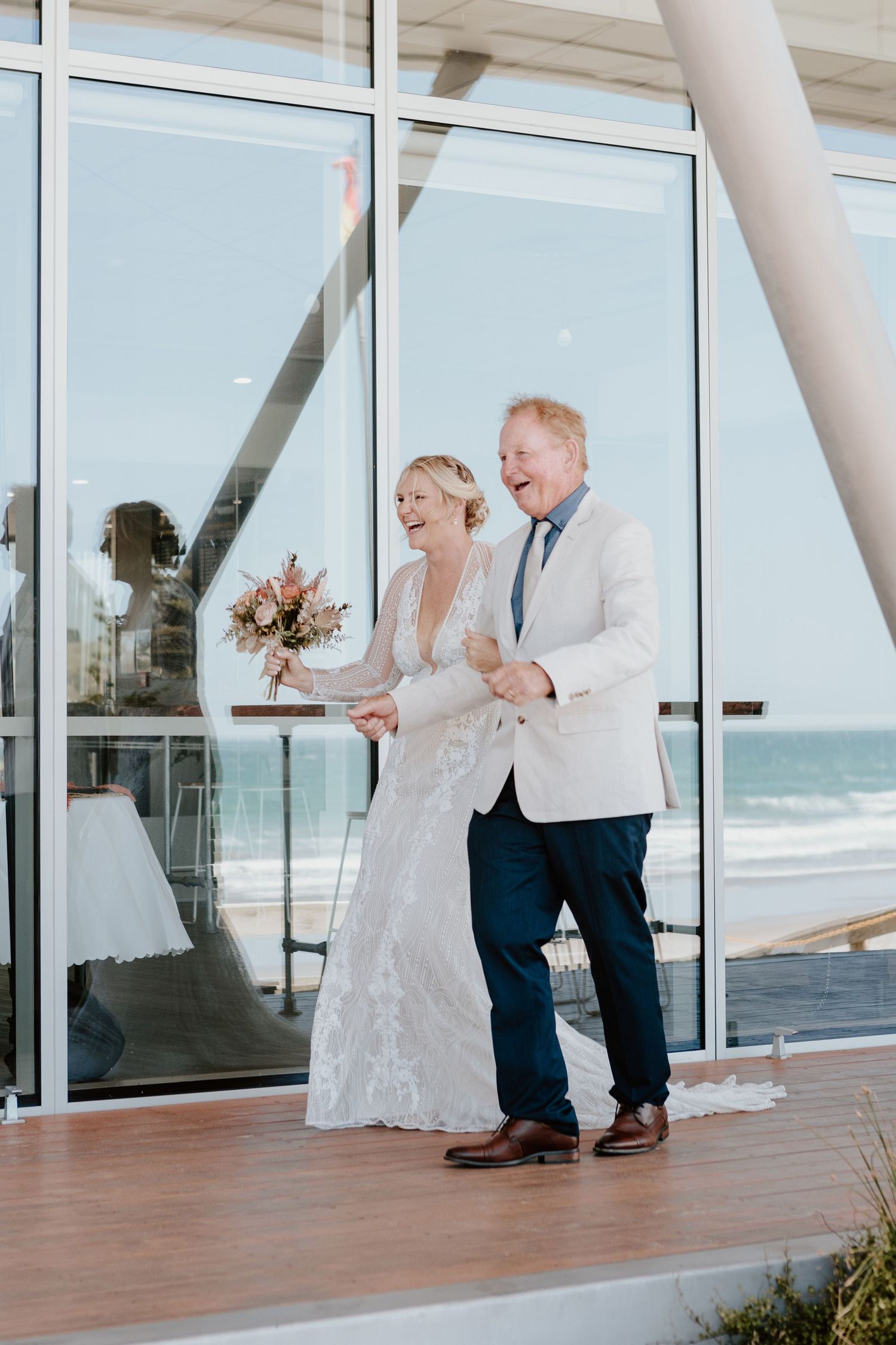 A joyful wedding moment captured at a beachside venue with glass windows and ocean views in the background.