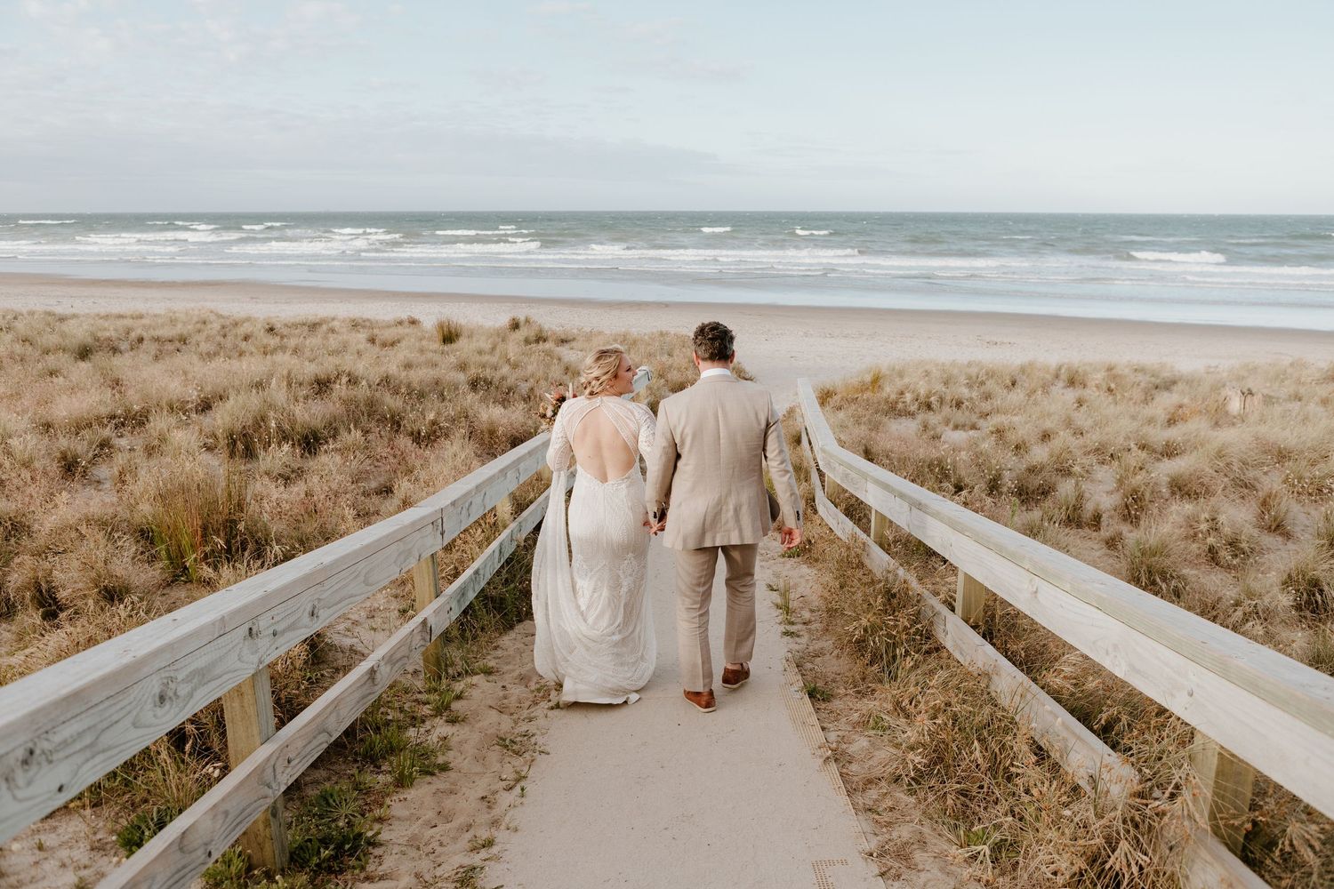 A couple walks down a wooden boardwalk toward the ocean on their wedding day at a coastal beach.