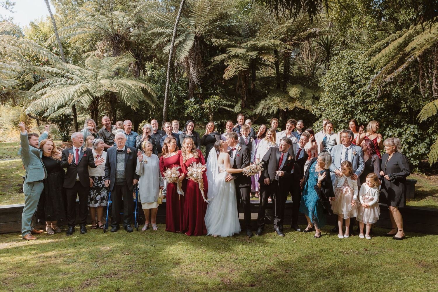 Full wedding party and guests pose for formal group photo in garden setting.